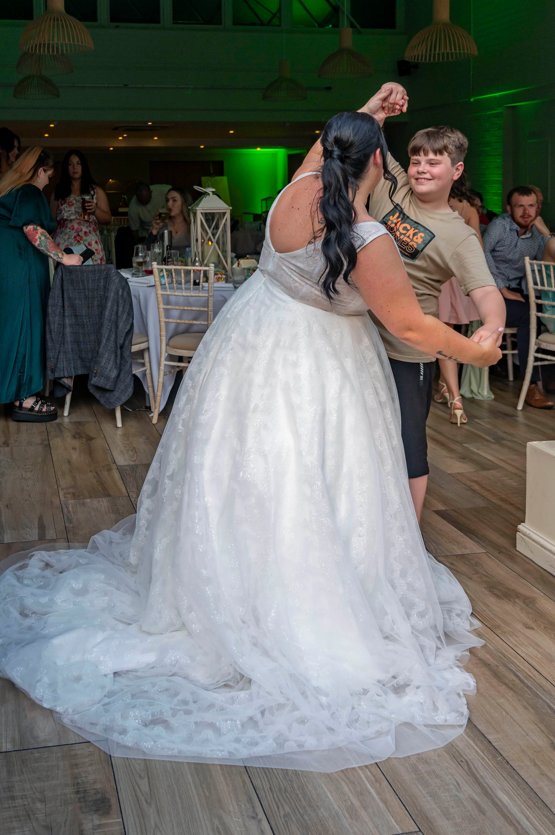 A bride in a sparkling white a-line gown dances joyfully on a wooden floor with a young guest holding hands.