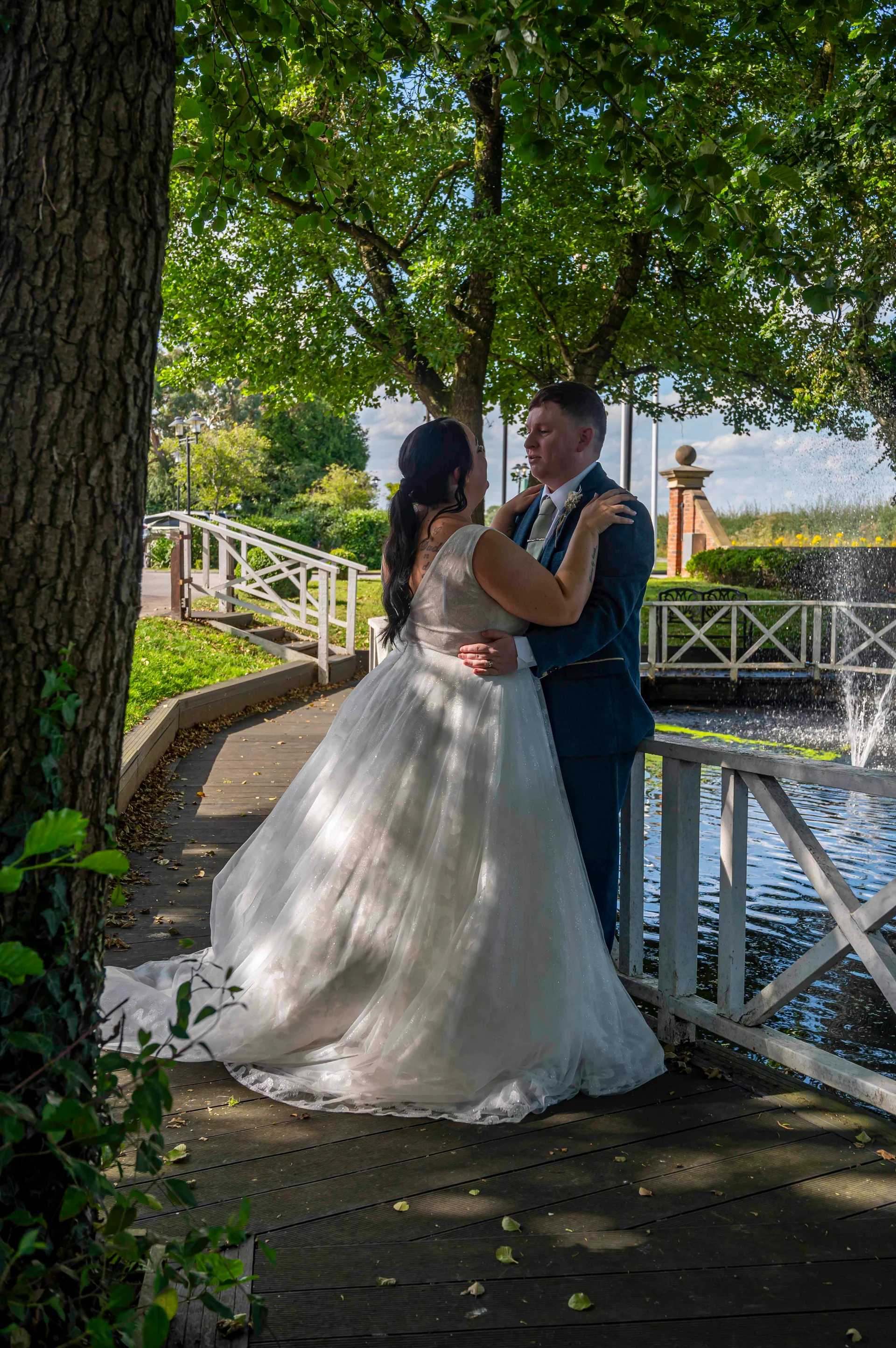 A newlywed couple embraces on a wooden bridge by an ornamental lake and fountain during a sunny summer wedding.