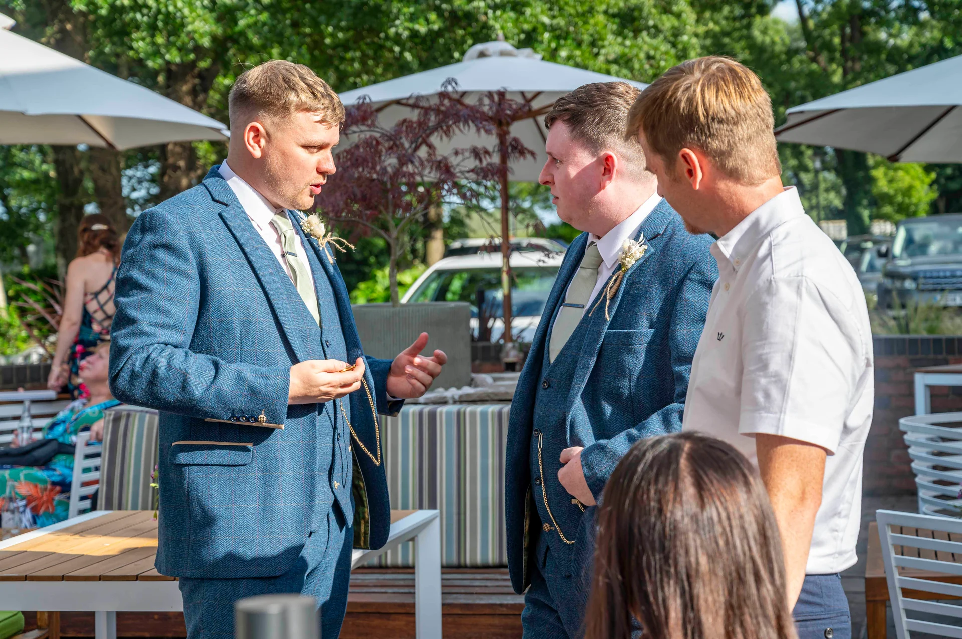 Three men in matching blue tweed suits and a guest in a white shirt chat outdoors in the sunshine near patio furniture and white parasols.