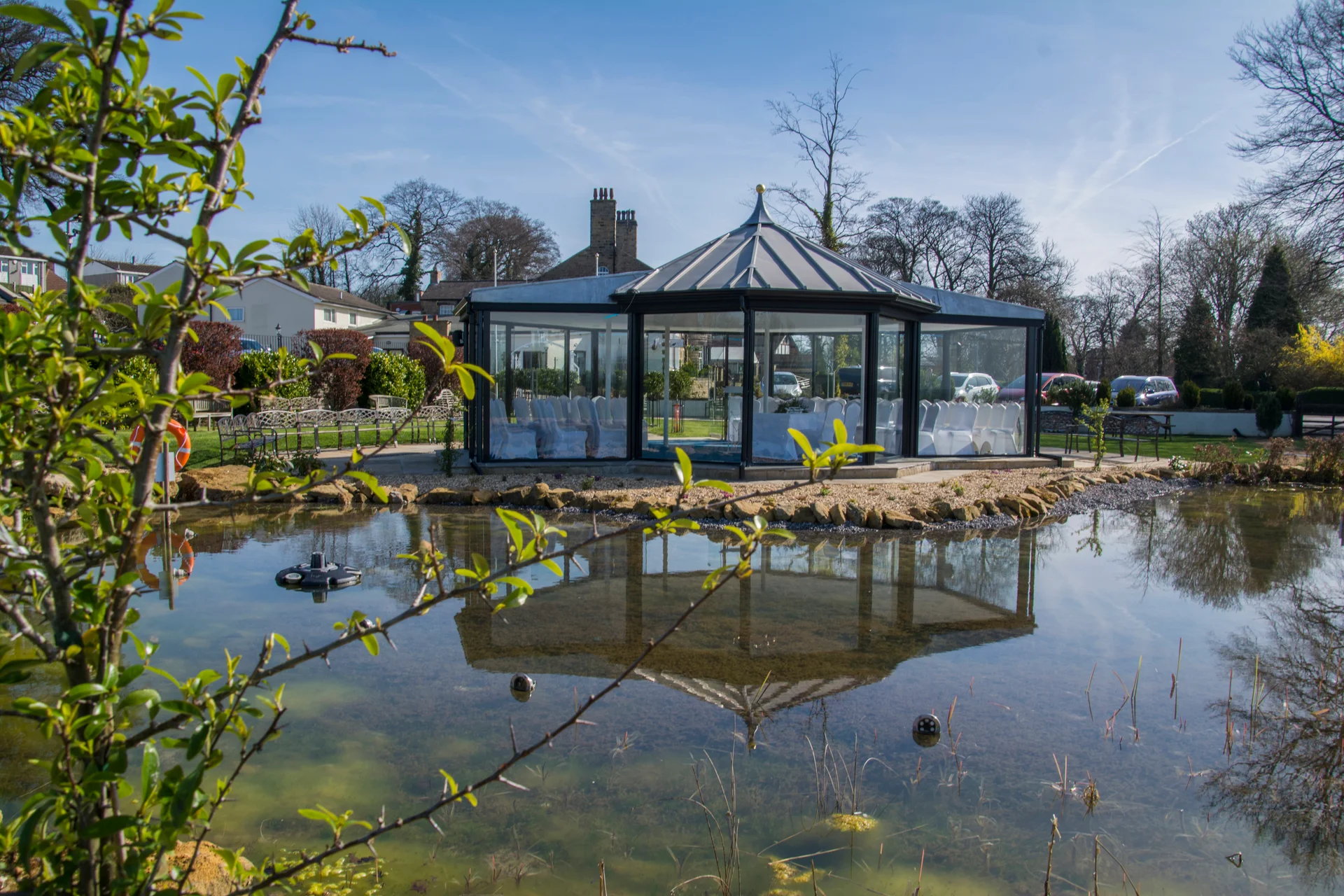 A modern glasshouse ceremony venue sits beside a tranquil lake on a bright, sunny summer day, reflecting clearly in the water.