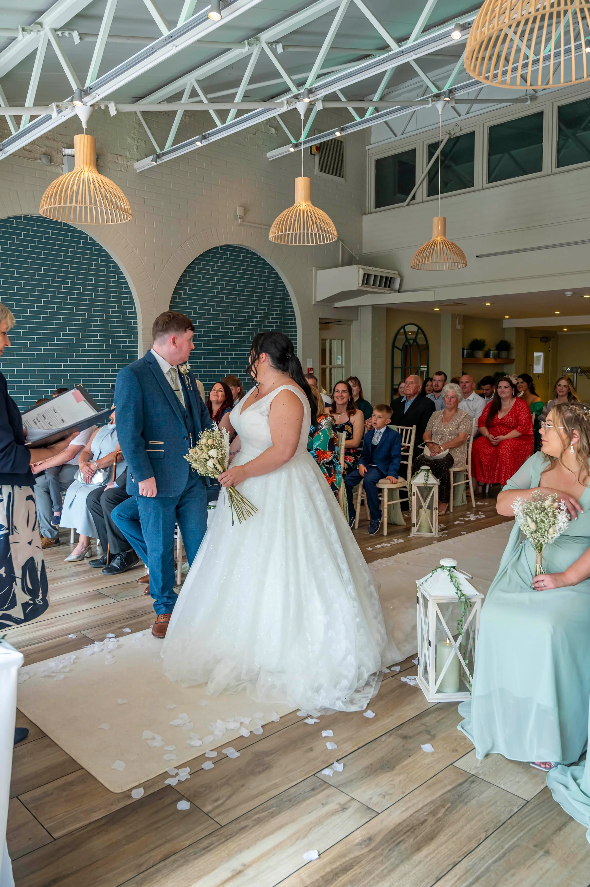 A bride in a white A-line gown and groom in a blue suit stand facing each other during their indoor wedding ceremony, surrounded by seated guests and lanterns along the aisle.