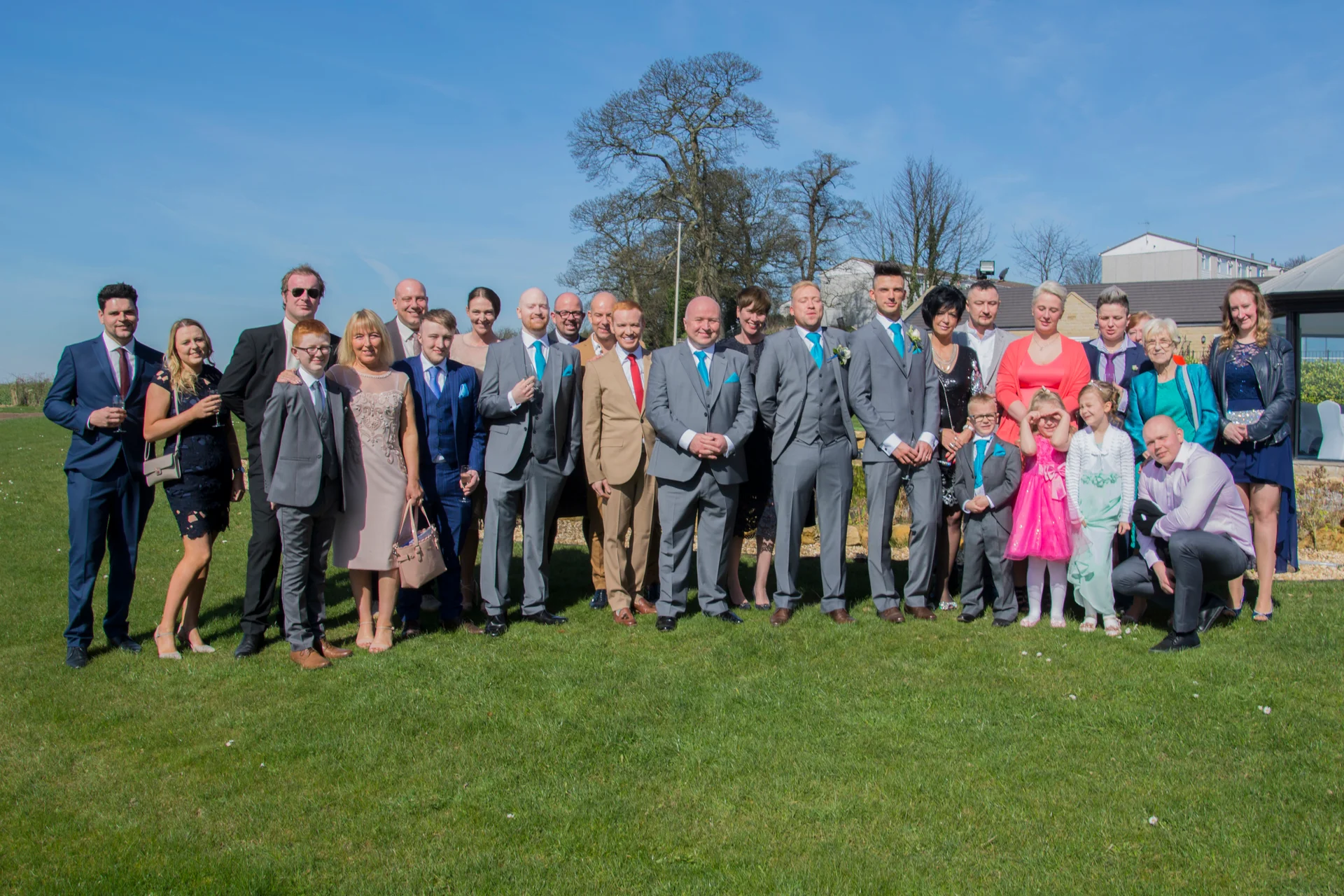A large group of wedding guests and family members pose together outdoors on a sunny green lawn under a clear blue sky.