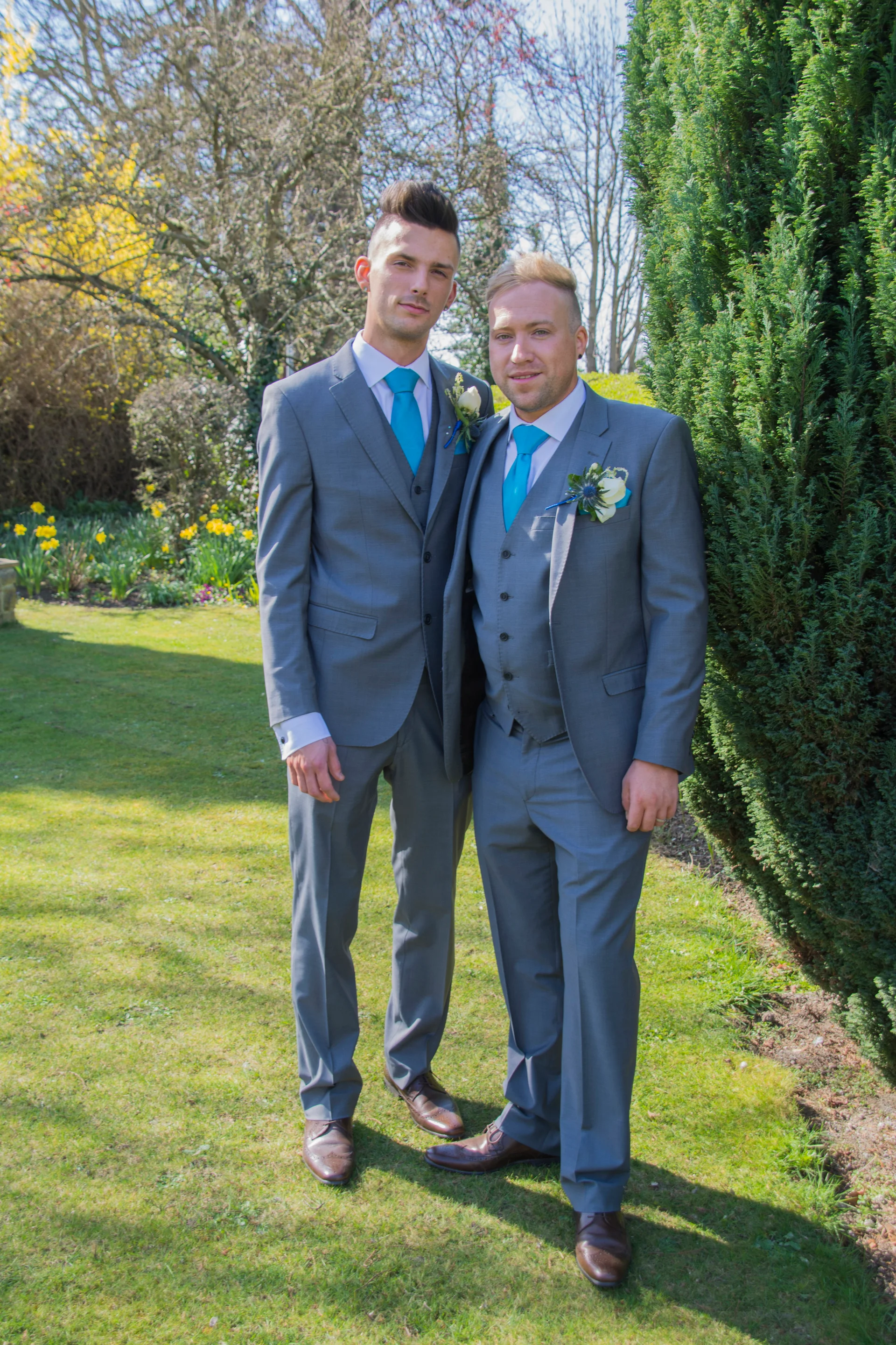 Two grooms in matching grey three-piece suits with bright blue ties and boutonnieres stand side-by-side in a manicured garden for a portrait.