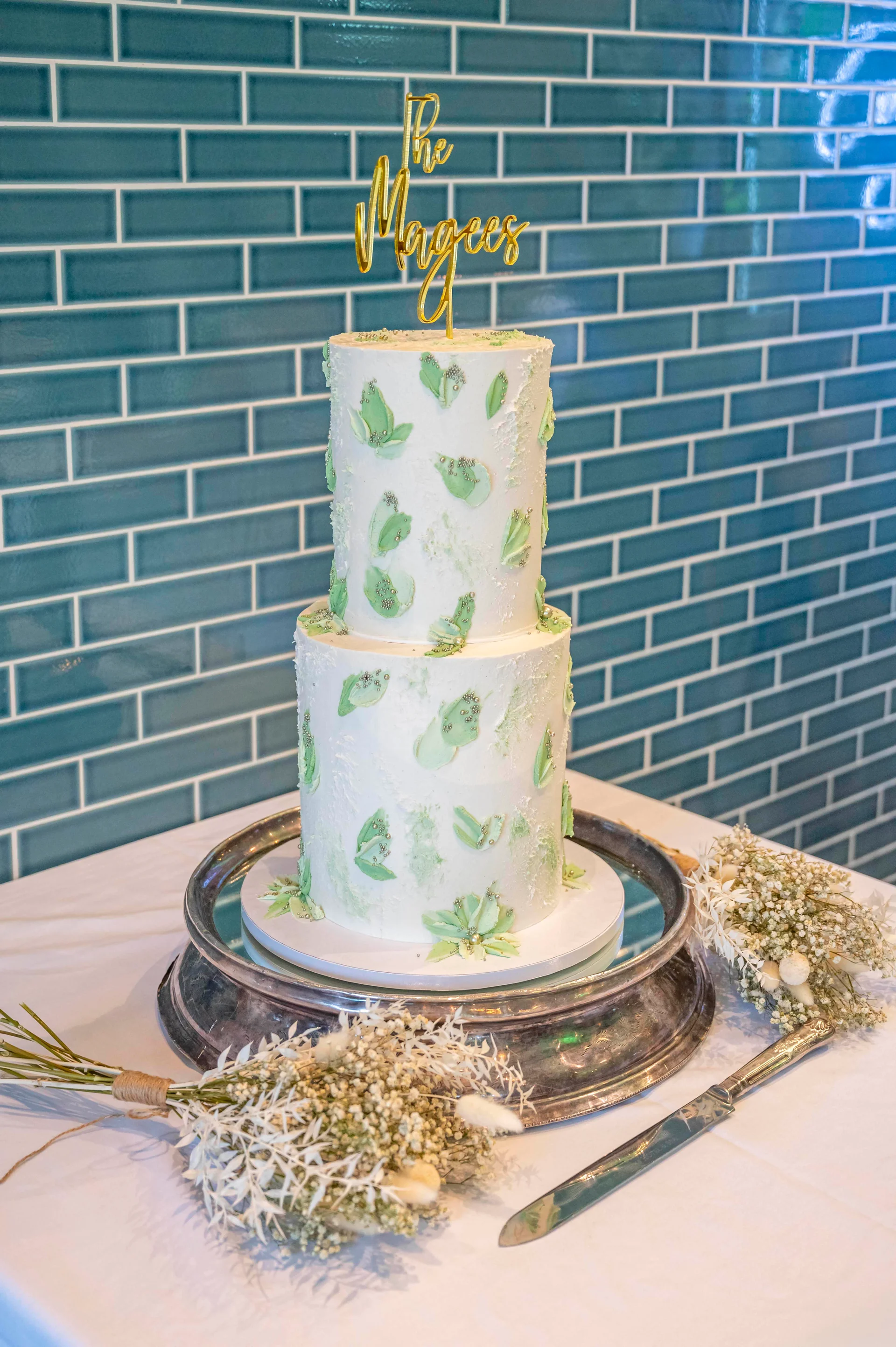 A two-tier white buttercream wedding cake decorated with sage green leaf accents and a gold acrylic topper sits on a silver cake stand against a teal tiled wall.