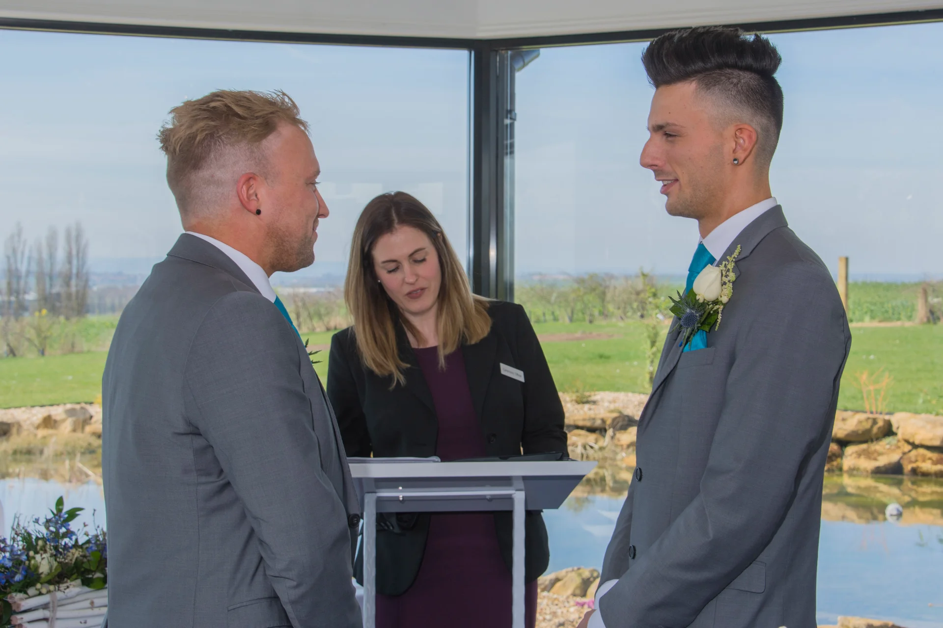 Two grooms in matching grey suits exchange vows during an indoor civil ceremony framed by large windows overlooking a scenic landscape.