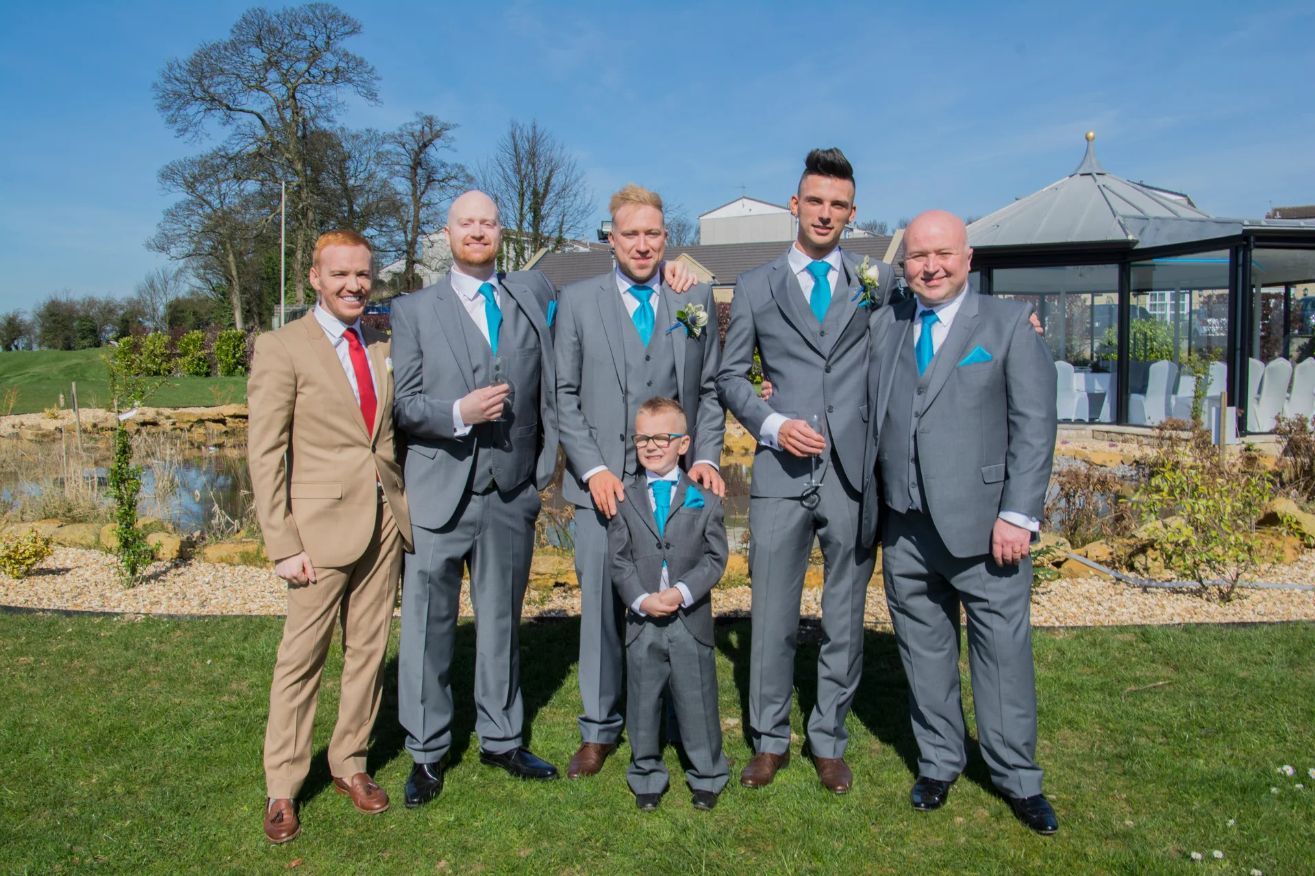 A groom and his groomsmen in grey and tan suits with bright blue ties pose outdoors on a sunny day.