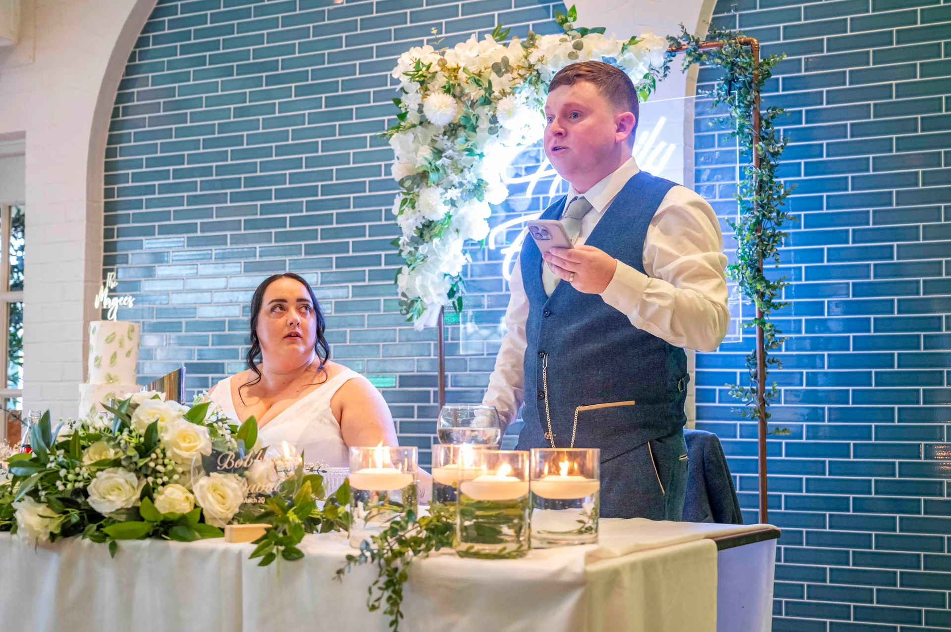 A groom in a blue tweed waistcoat delivers a speech while holding his phone at the top table, as a bride looks up attentively in front of a blue tiled wall and floral arch.
