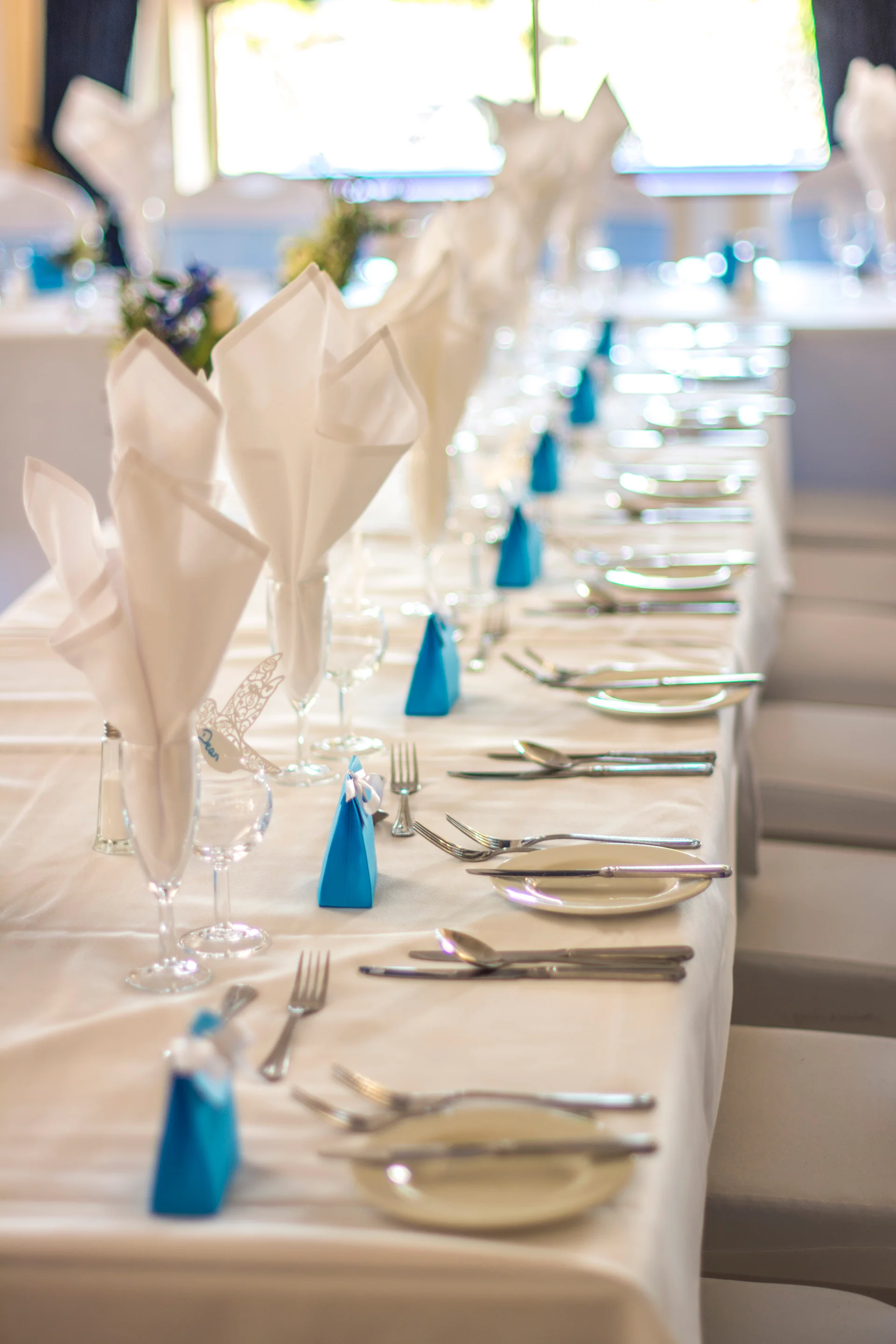 An elegantly set long banquet table features crisp white linens, neatly folded napkins, and vibrant blue favor boxes in a bright, light-filled venue.