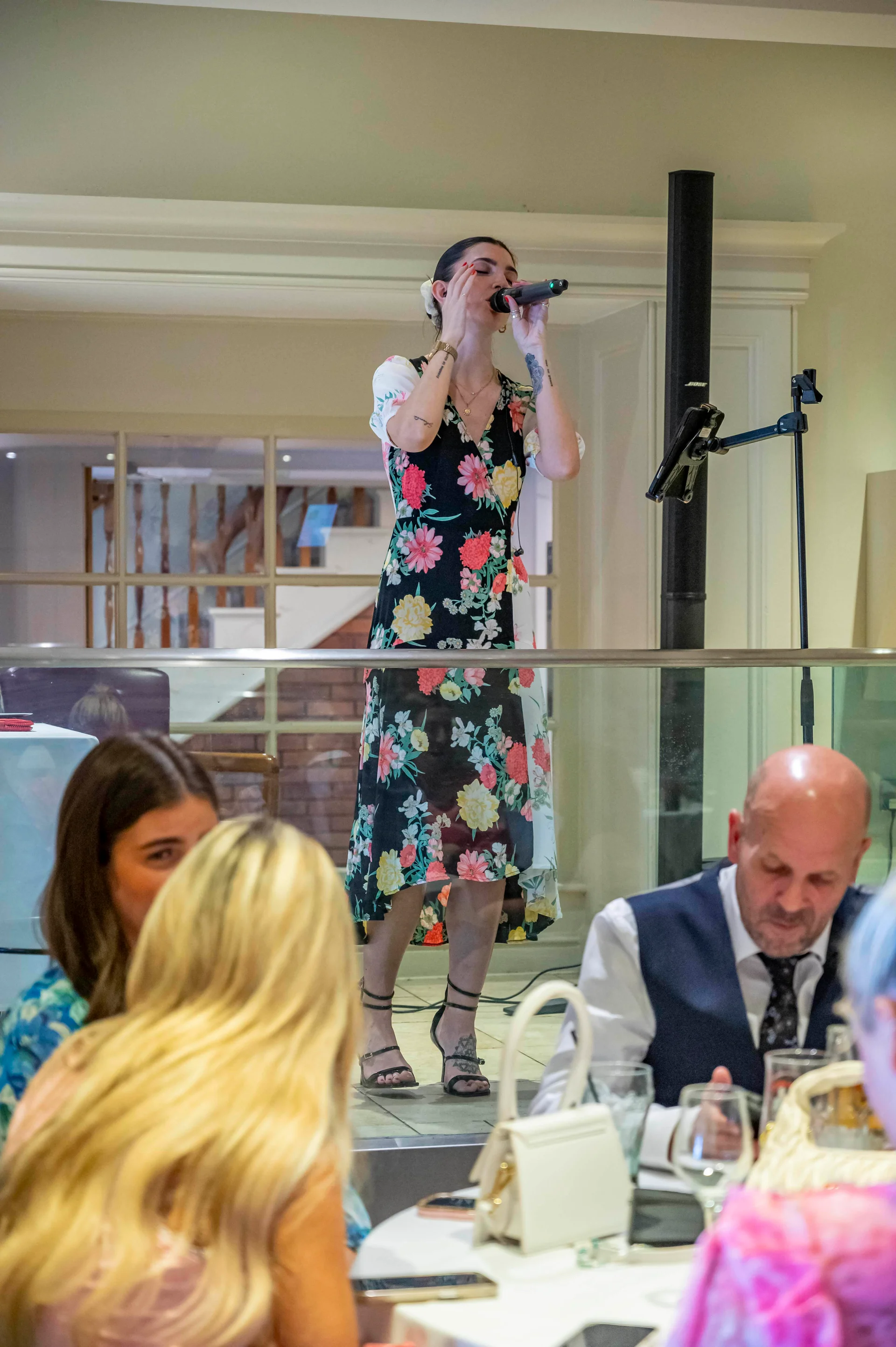 A female wedding singer performs with a microphone in front of seated guests during a wedding reception.