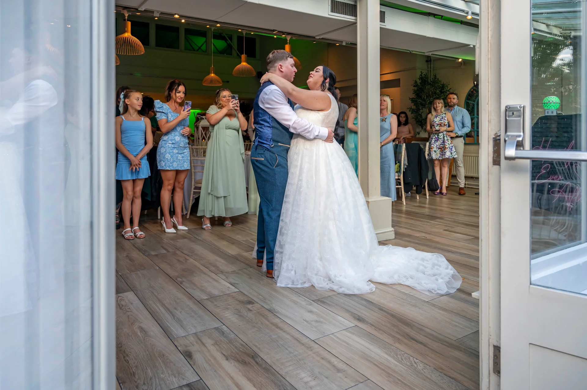 A newlywed couple shares their first dance in a light-filled reception venue while family and friends watch and capture the moment on their phones.