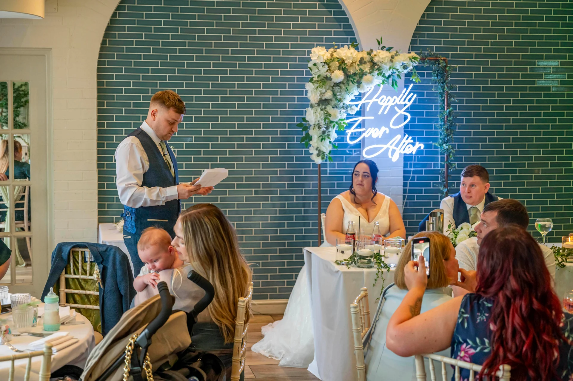 A wedding guest stands to deliver a speech while seated guests listen and look on during the wedding reception.