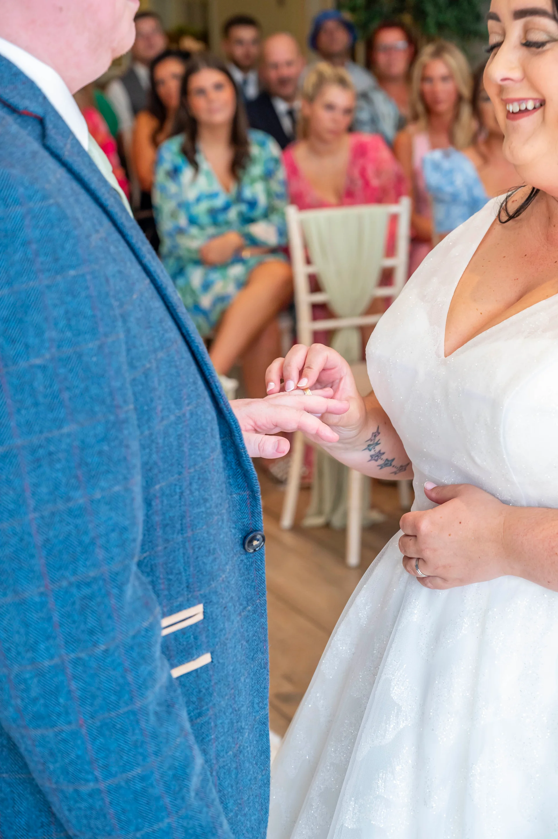 A smiling bride wearing a white v-neck gown places a wedding ring onto the groom's finger during the ceremony while guests watch in the background.
