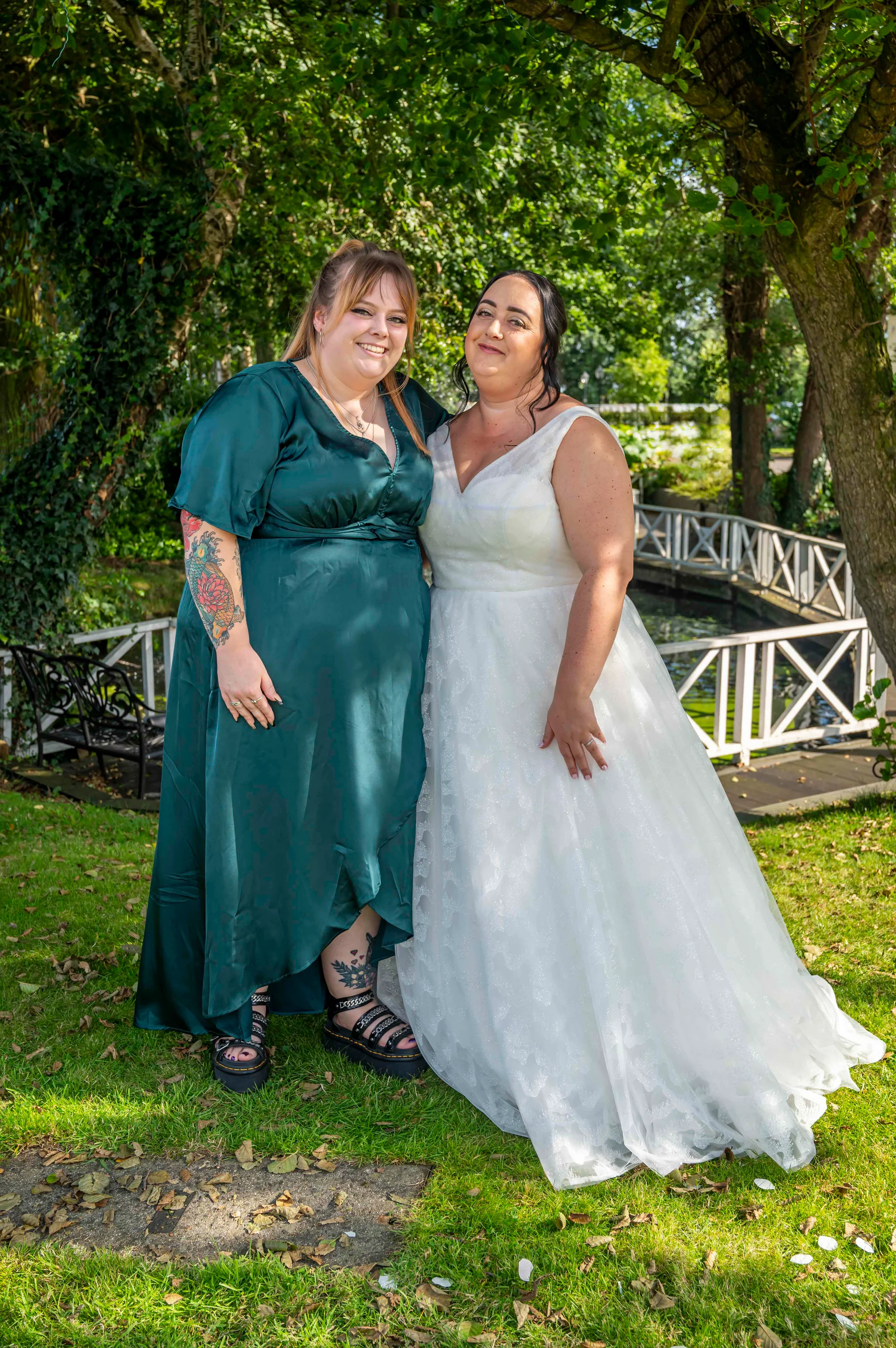 Two smiling brides stand side by side on a lush green lawn in an outdoor garden setting, one wearing a vibrant emerald green dress and the other in a classic white wedding gown.