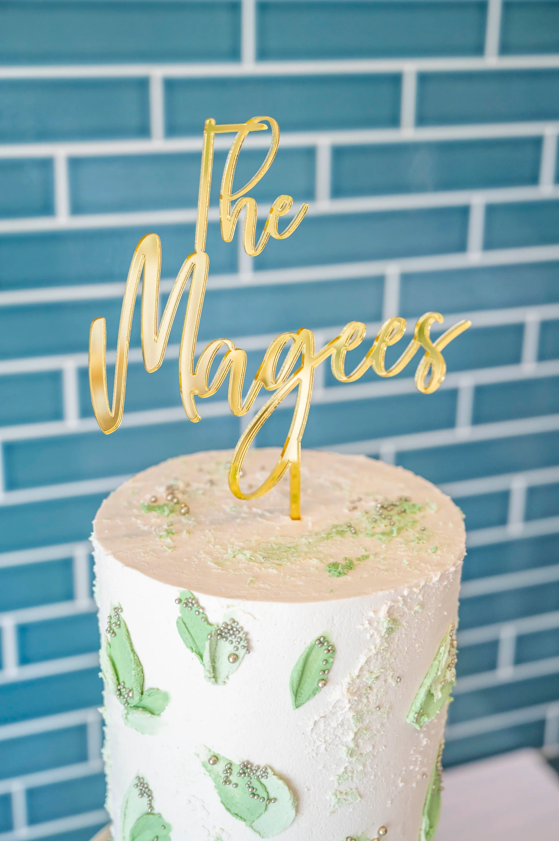 A close-up shot of a single wedding cake decorated with textured white buttercream and delicate green leaf details, topped with a shiny gold acrylic cake topper against a blue tiled wall.