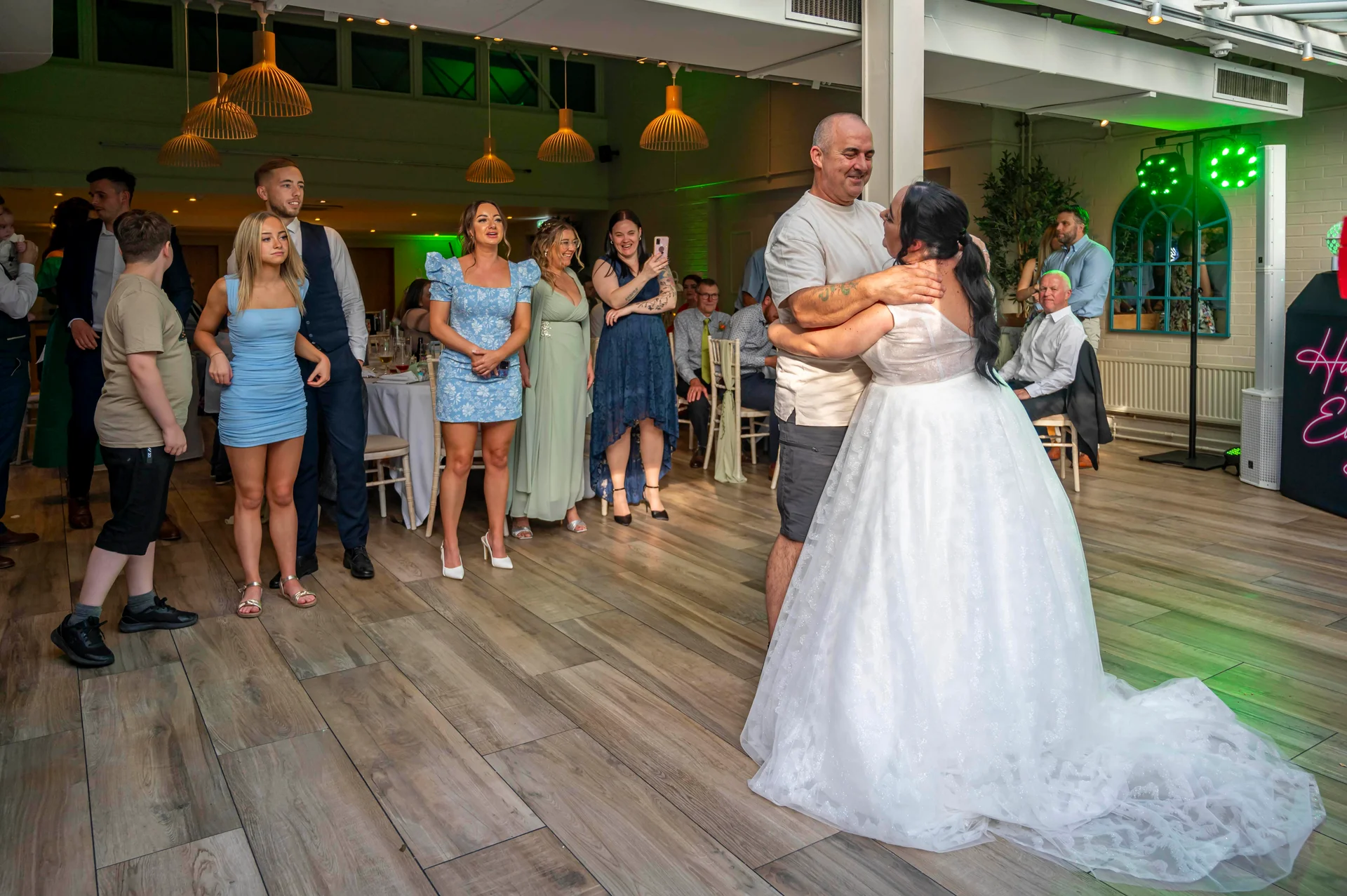 A newlywed couple shares their first dance on a wooden floor inside a bright venue while guests watch and smile.