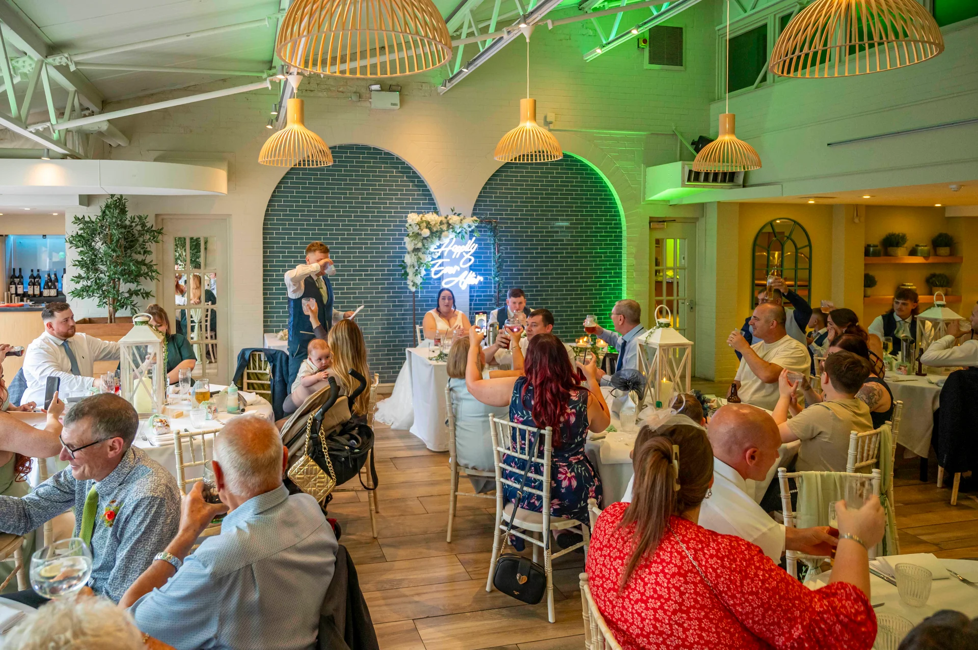 Wedding guests enjoy speeches and raise glasses during the wedding breakfast in a modern restaurant venue featuring wooden pendant lighting and green tiled arches.