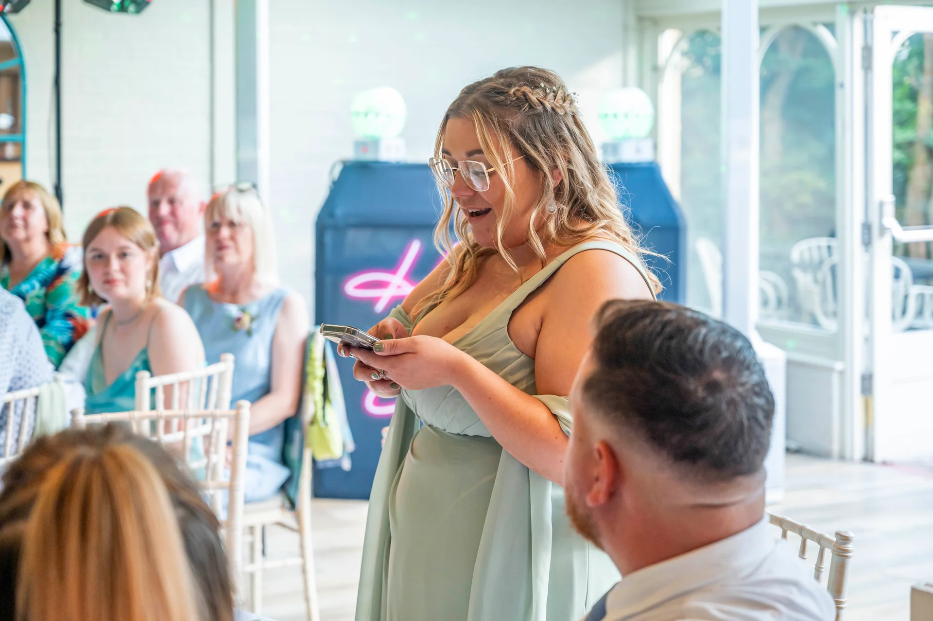 A guest wearing a sage green dress and glasses looks down at her phone with an excited expression during wedding speeches.