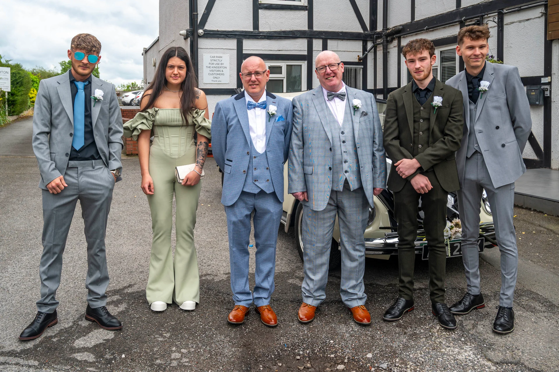 A diverse wedding party and guests pose outside a traditional venue next to a classic car in tailored grey, checked, and olive suits.