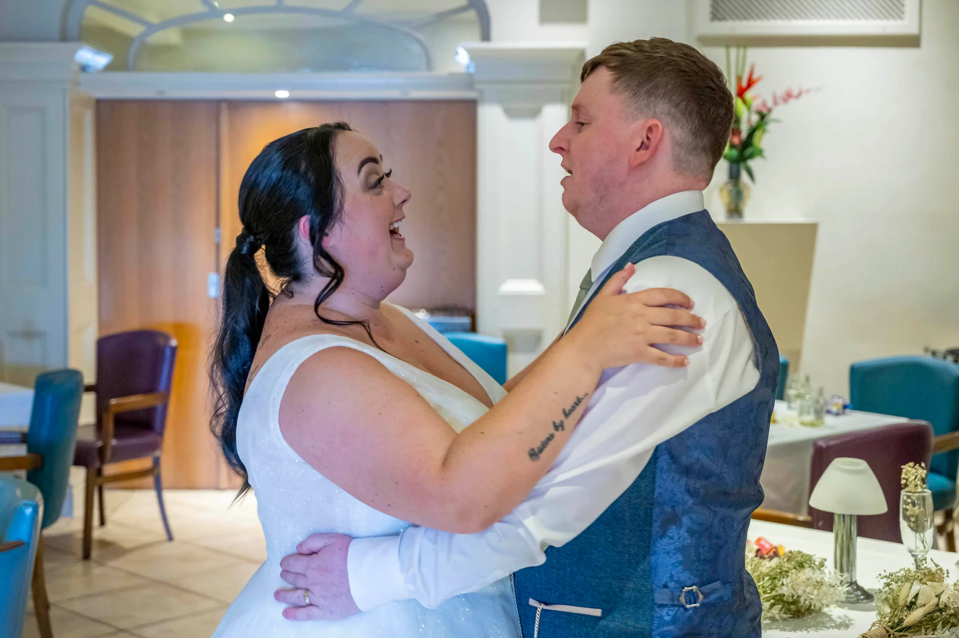 A newlywed couple shares a joyful and laughing moment during their wedding reception in an indoor venue.