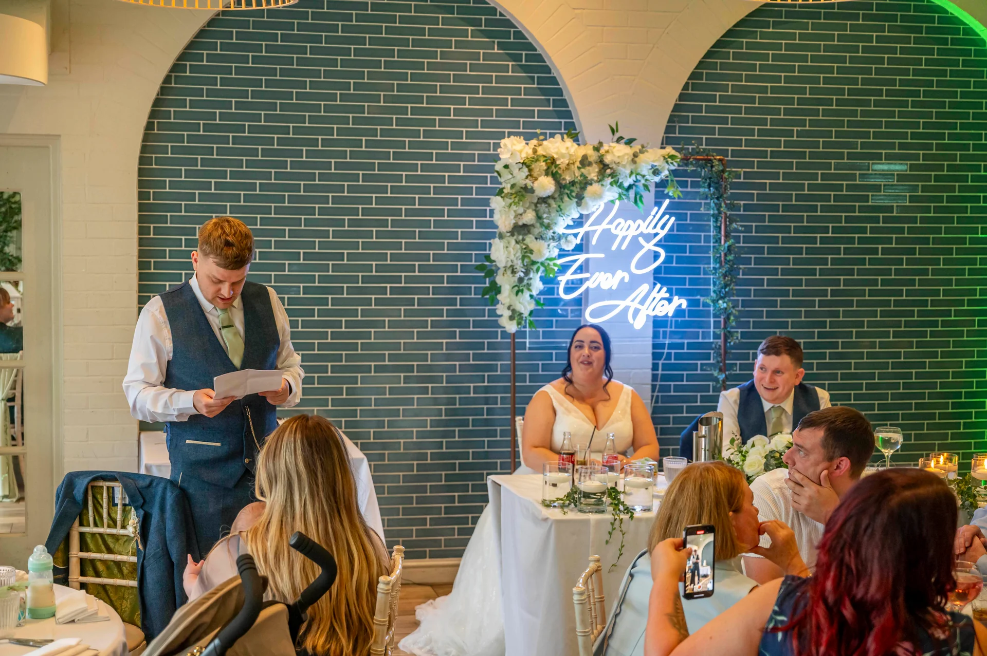 A groom delivers his wedding speech at the top table in front of a teal tiled wall and illuminated neon sign, with the bride and guests listening closely.