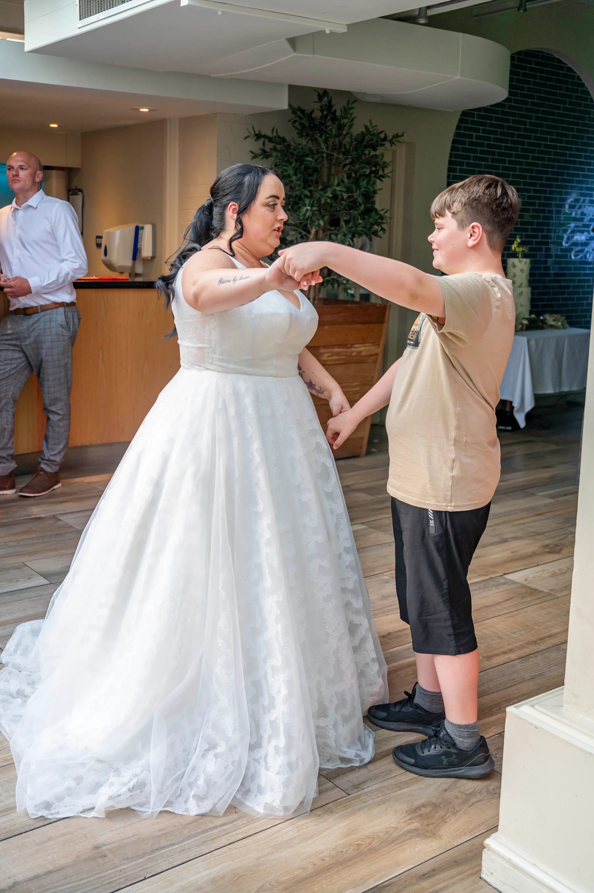A bride in a white tulle a-line gown holding hands and dancing with a young boy inside a modern venue.