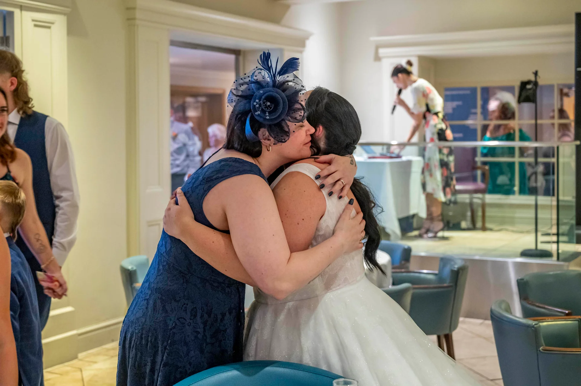 A wedding guest wearing a navy blue fascinator embraces the bride in a warm, emotional hug inside a venue reception room.
