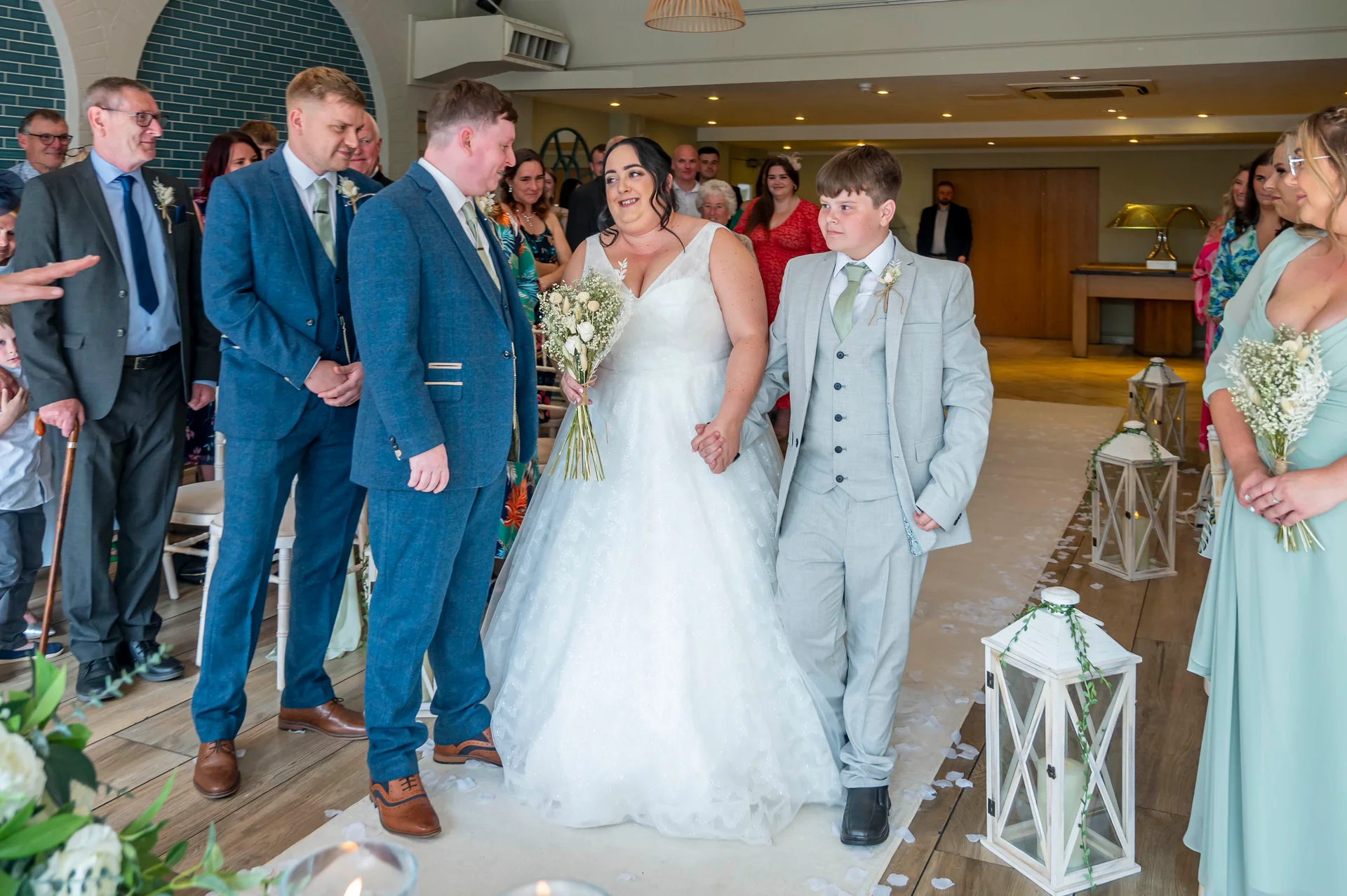 A smiling bride and groom walk down the aisle together accompanied by a page boy holding their hands during a light-filled indoor ceremony.