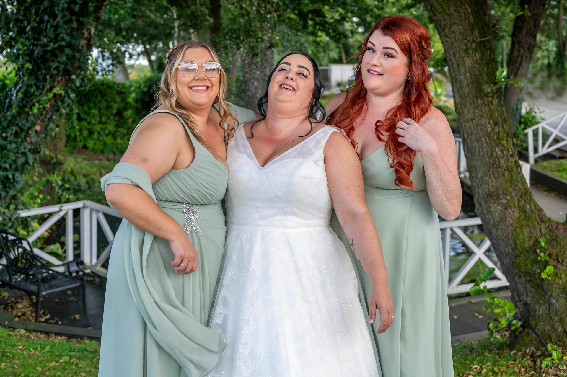A joyful bride in a white A-line gown laughs heartily outdoors alongside two bridesmaids wearing sage green chiffon dresses near a picturesque lakeside setting.