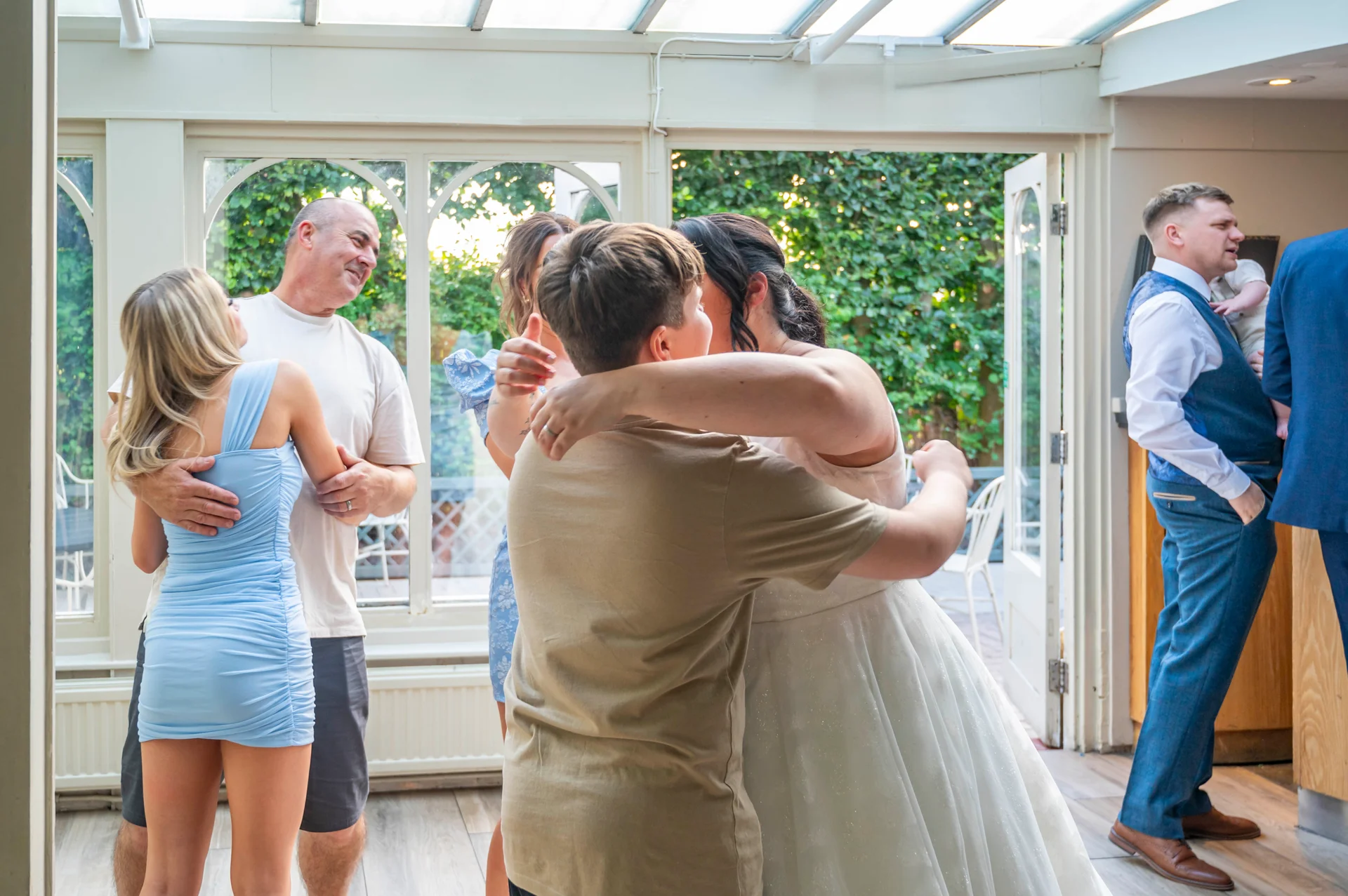 Guests and family mingle warmly in a bright conservatory during a wedding reception, sharing hugs and candid conversations.