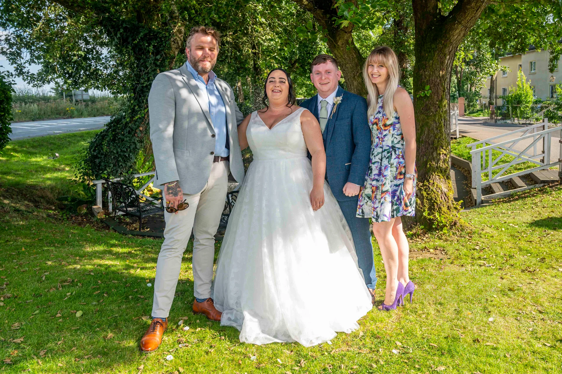 A bride in a white tulle gown stands smiling alongside a groom in a blue suit, flanked by two guests outdoors in a lush, tree-lined garden.