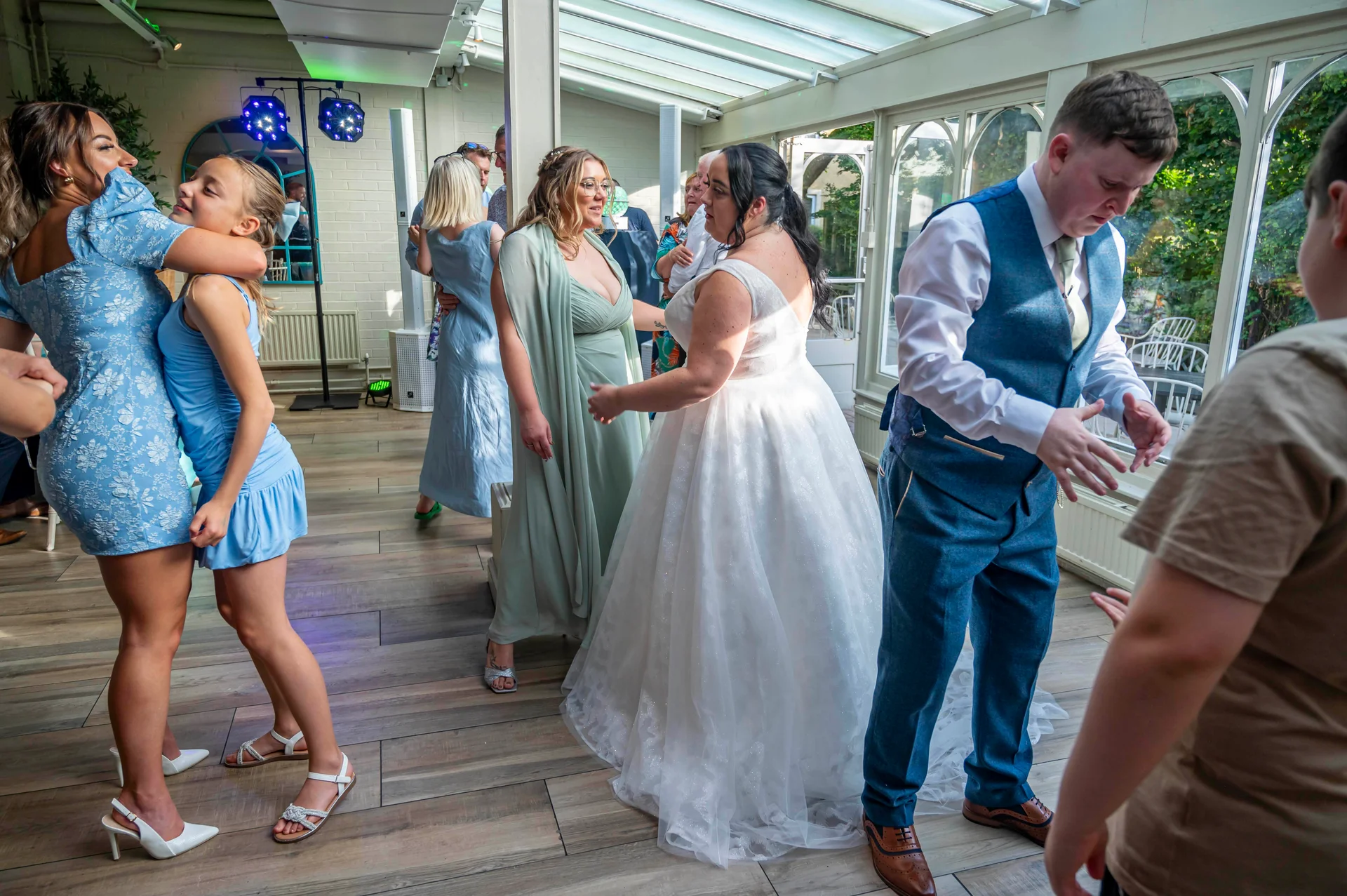 Guests and newlyweds enjoy dancing and celebrating on the wooden floor of a sunlit conservatory venue.
