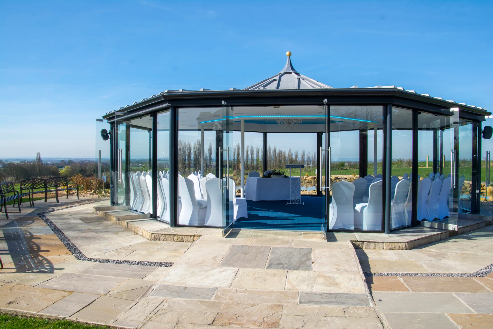 A modern glass gazebo wedding venue stands on a paved terrace under a clear blue sky, prepared with rows of white chairs.