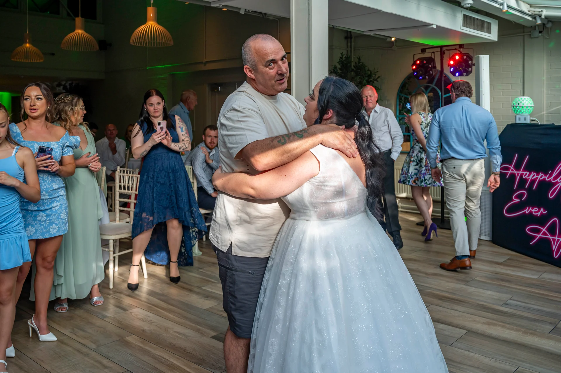 A bride in a sparkling A-line wedding dress dances with a guest on the wooden dance floor while friends and family look on smiling.