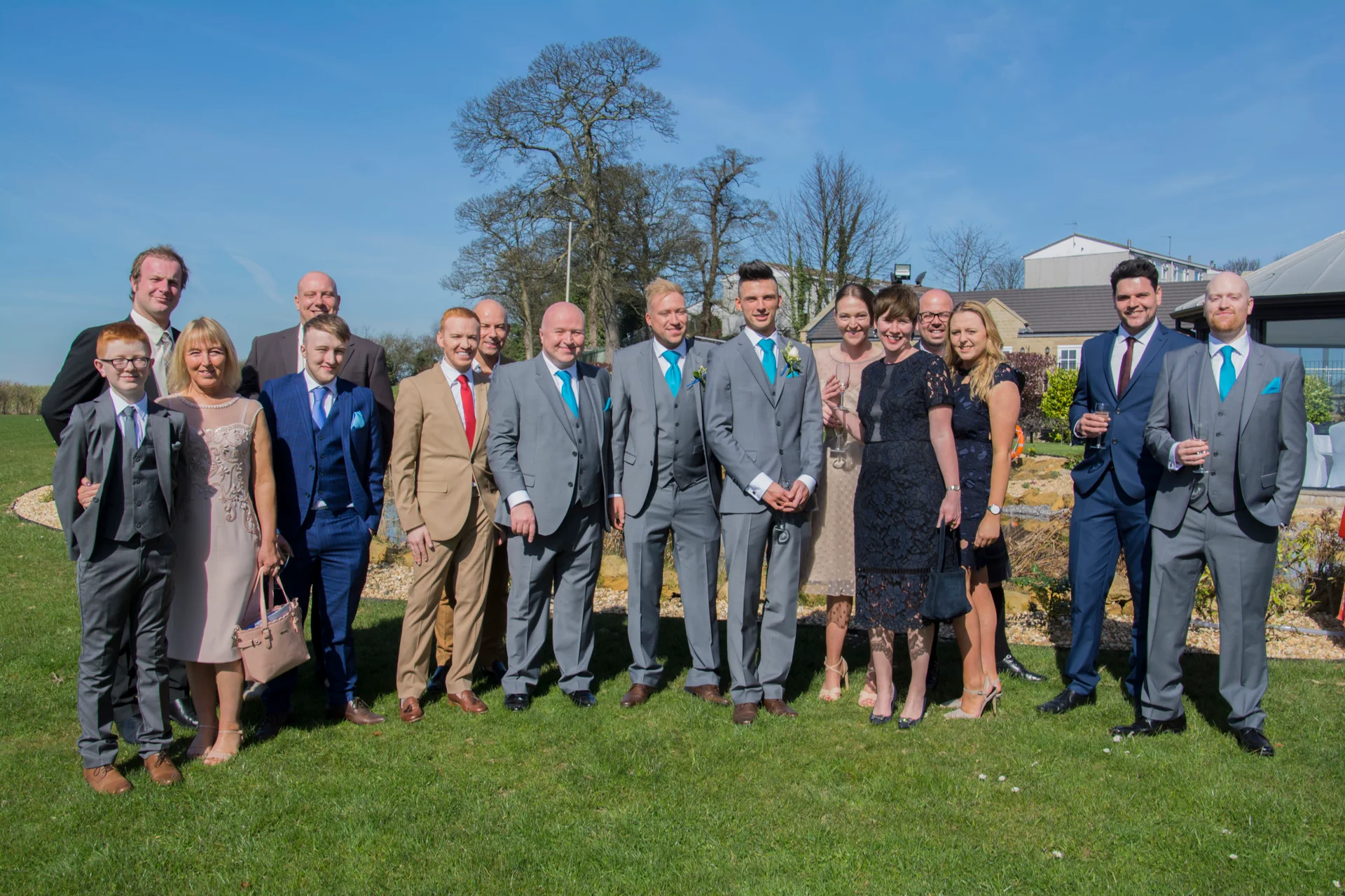 A large wedding party and family members pose together outdoors on a manicured lawn under a clear blue sky on a sunny day.