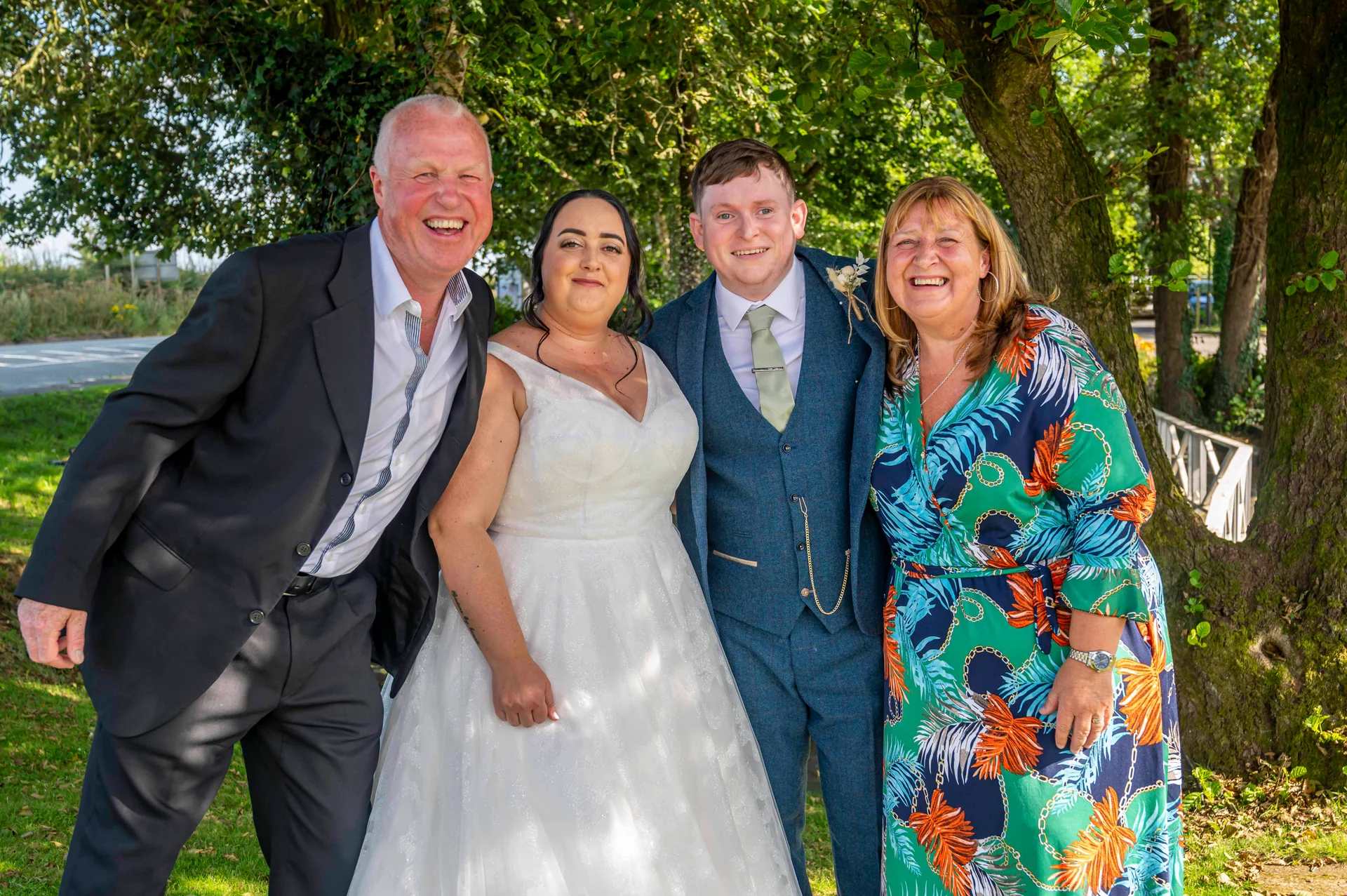 The bride and groom pose happily outdoors with their parents in front of lush green trees on a sunny wedding day.