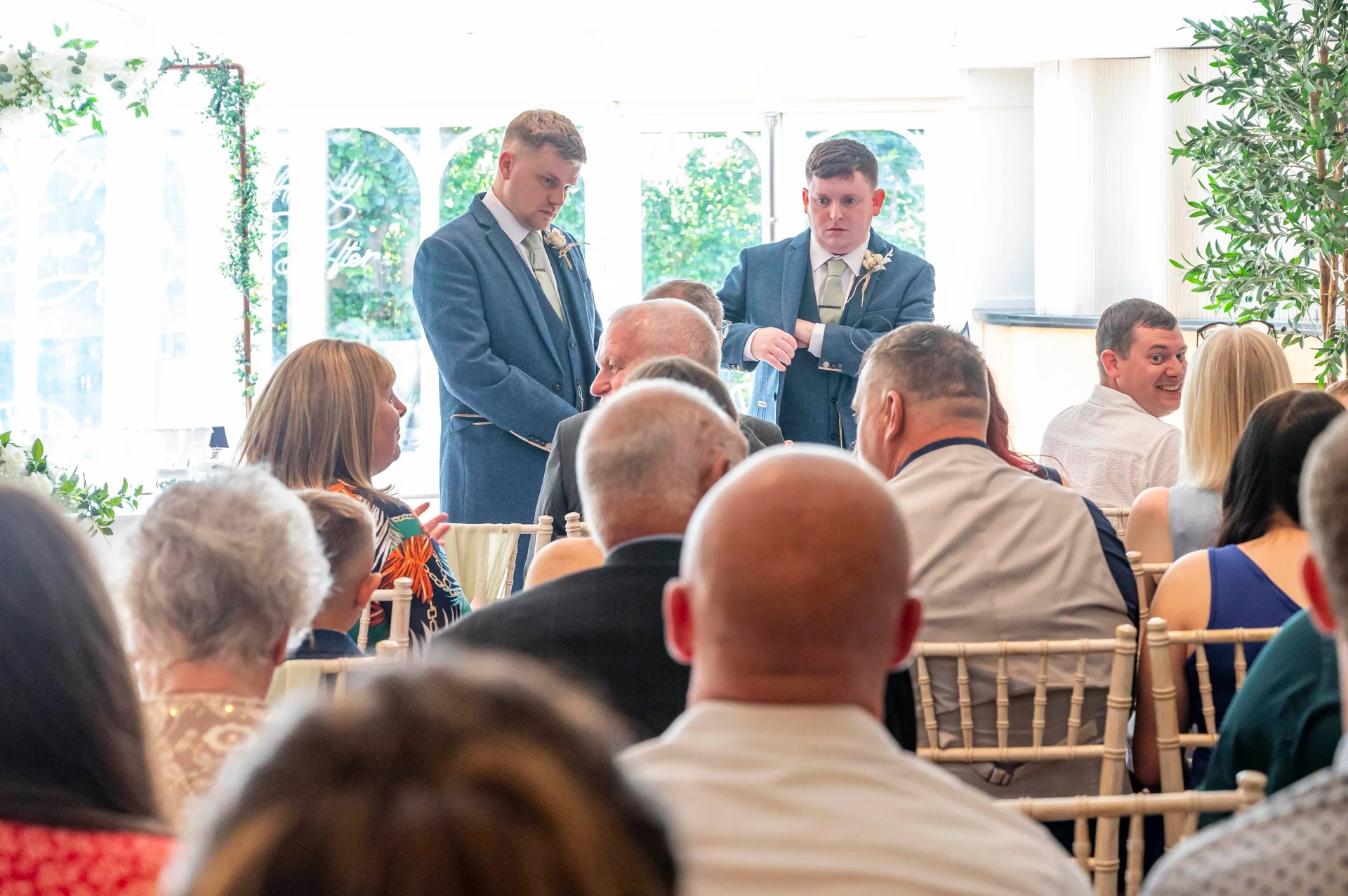 A candid wedding scene showing guests seated on chiavari chairs listening attentively while groomsmen stand at the front of the room.