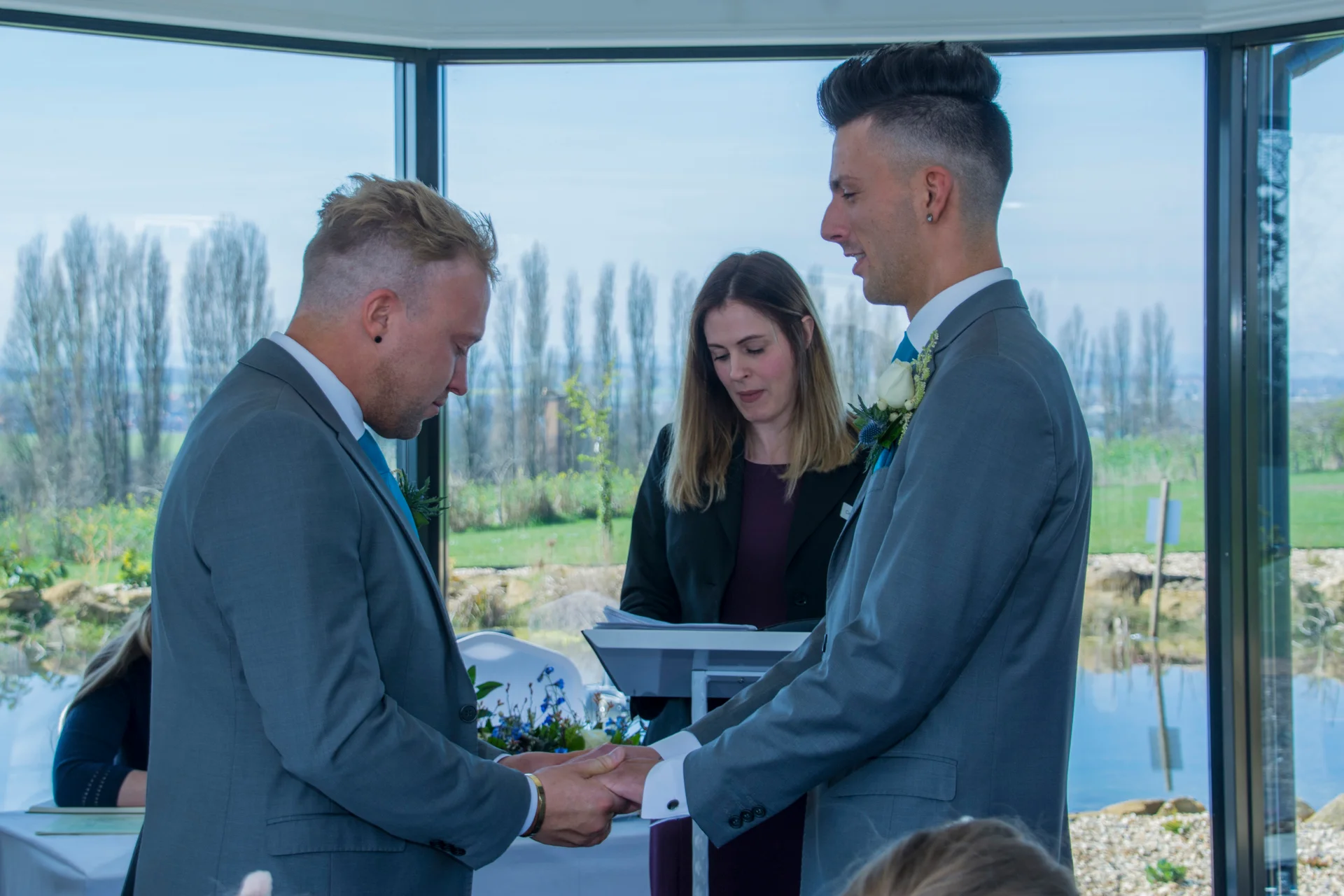 Two grooms stand holding hands during their civil ceremony inside a light-filled venue with large glass windows overlooking a landscape.