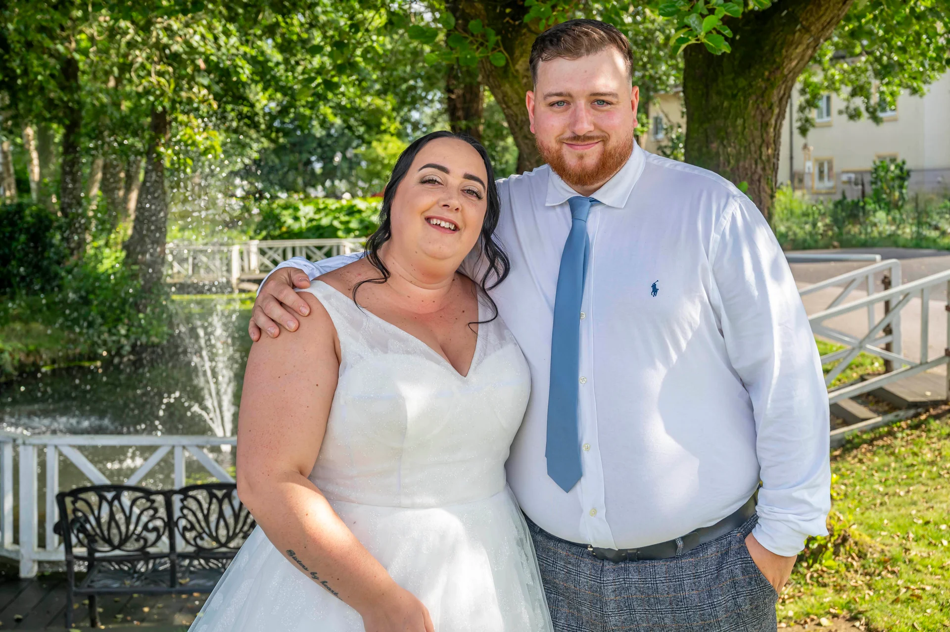 A smiling bride in a sparkly white V-neck gown and groom in a white shirt and blue tie pose outdoors by a fountain and lake.