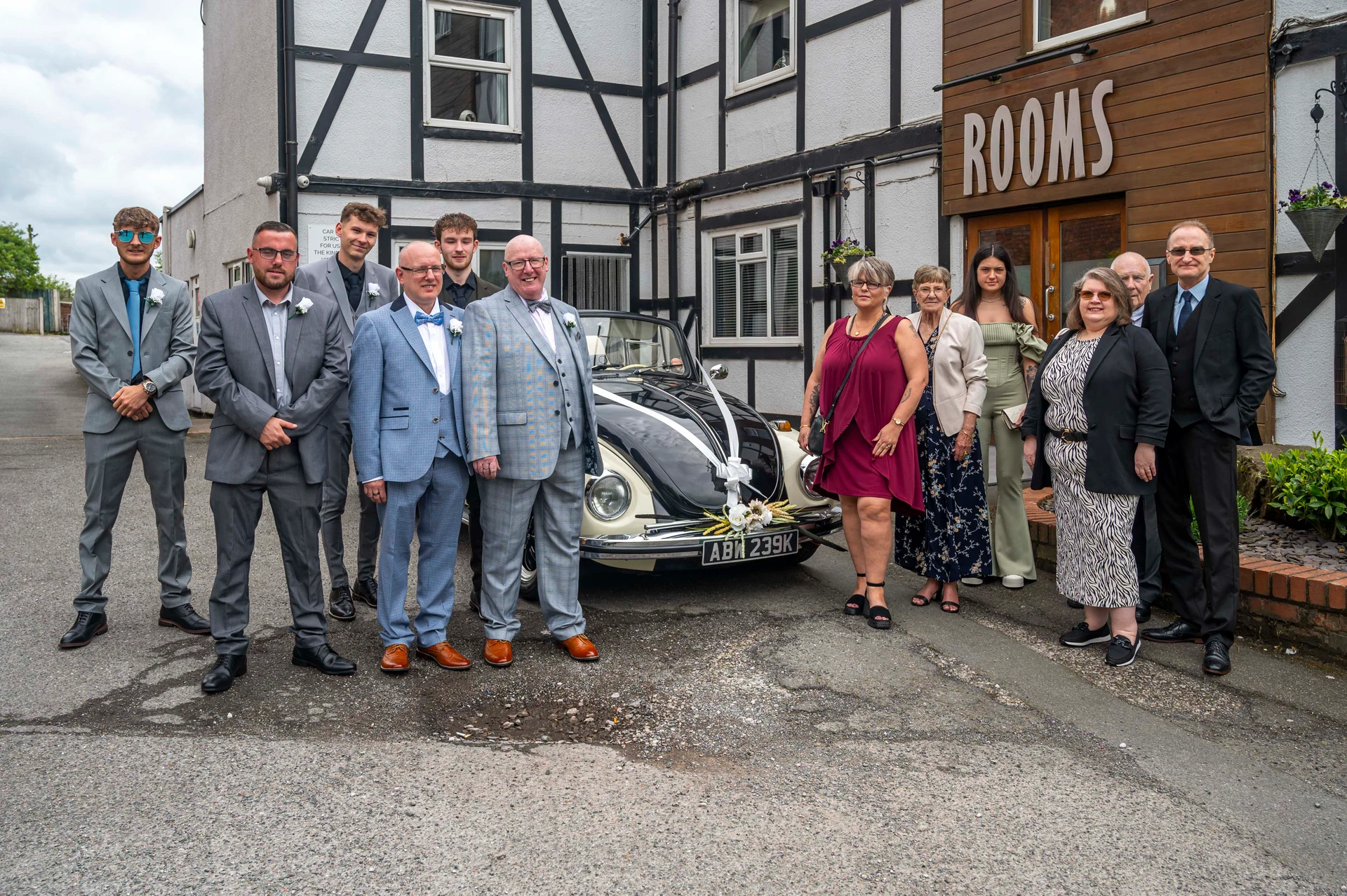 A wedding group poses together outside a venue next to a decorated vintage Volkswagen Beetle car on a bright day.