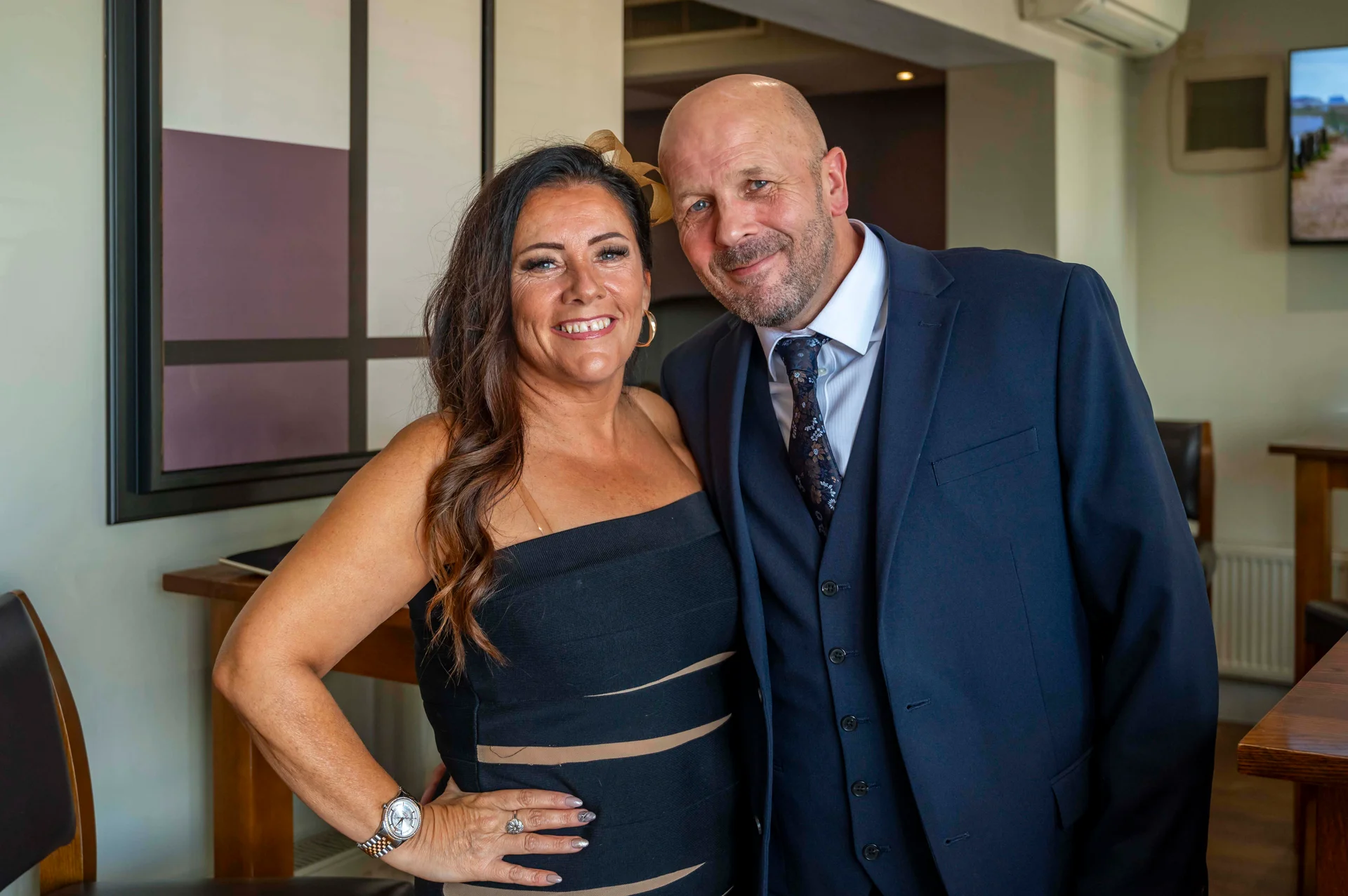 An older couple poses smiling together indoors, with the woman wearing a dark strapless dress and a fascinator, and the man wearing a navy three-piece suit with a patterned tie.