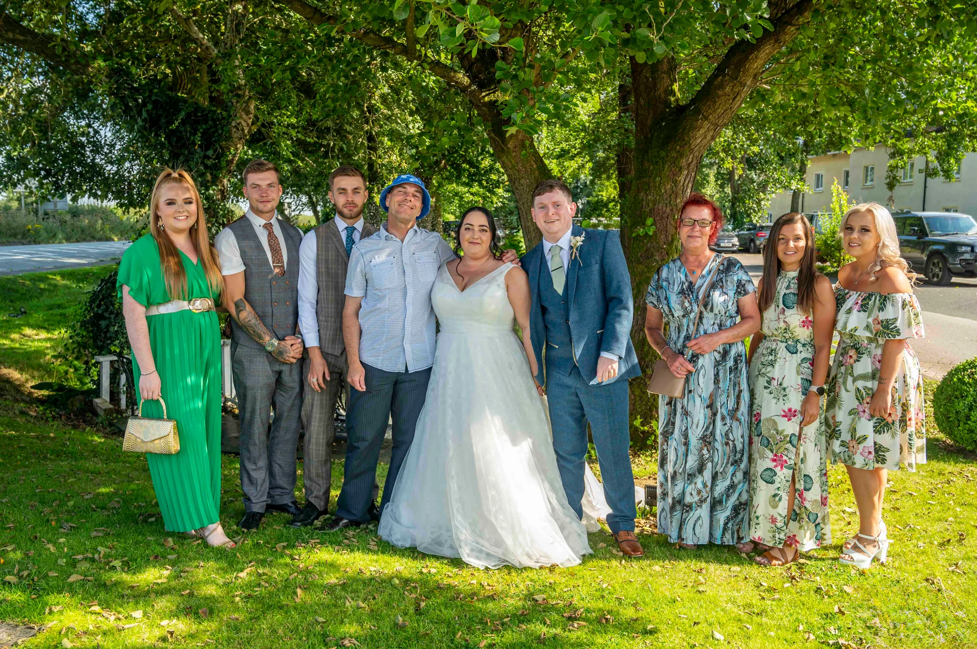 The bride and groom pose outdoors on a sunny lawn with wedding guests under the shade of a large green tree.