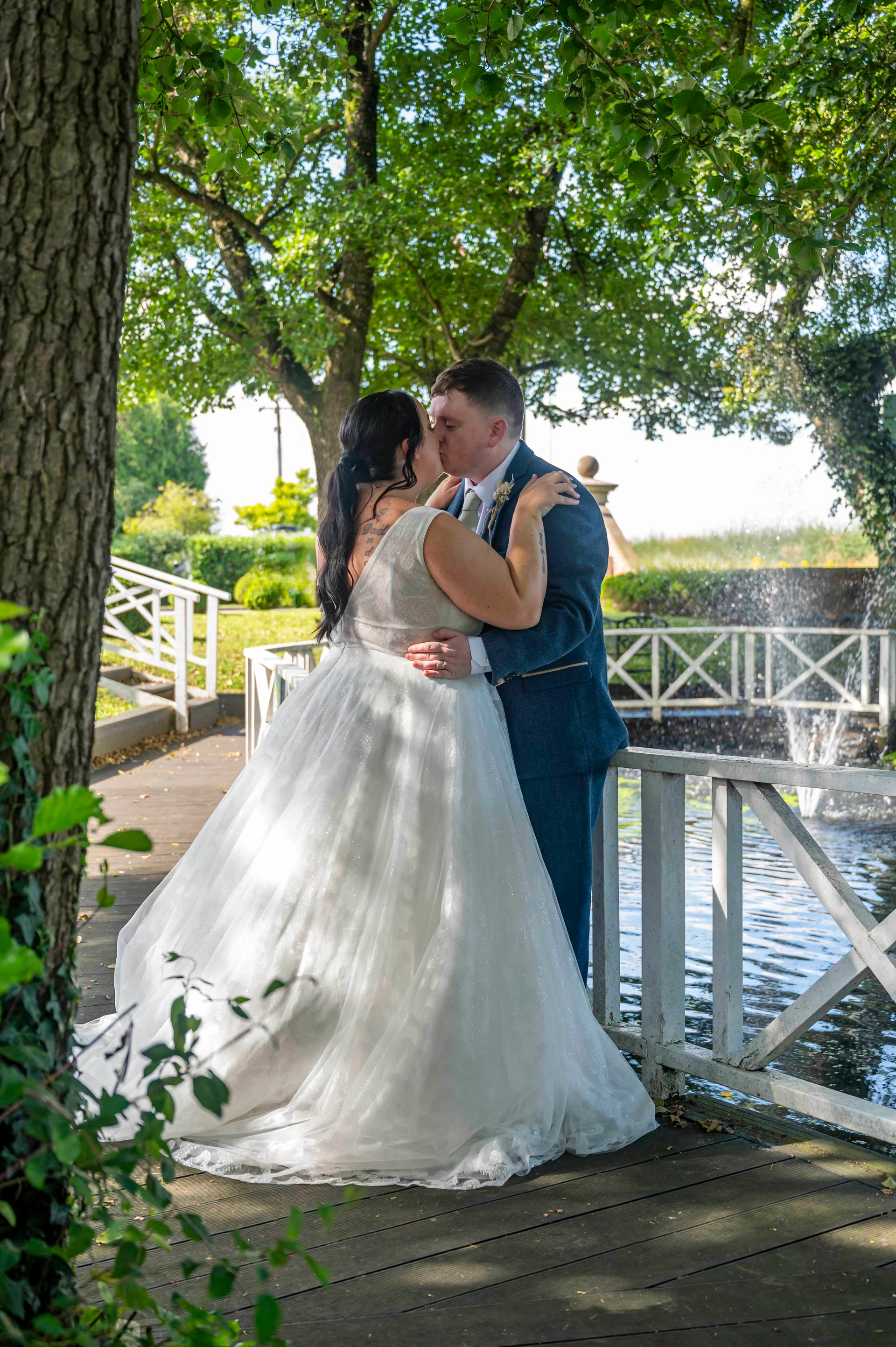 A bride in a glittering A-line wedding dress and a groom in a blue suit share a kiss on a wooden bridge by a fountain and lake.