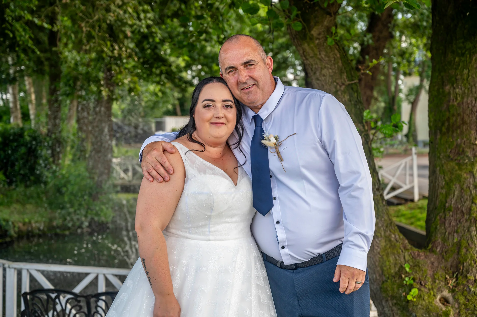 A proud father and bride pose together outdoors next to a mature tree and white wooden railing on her wedding day.