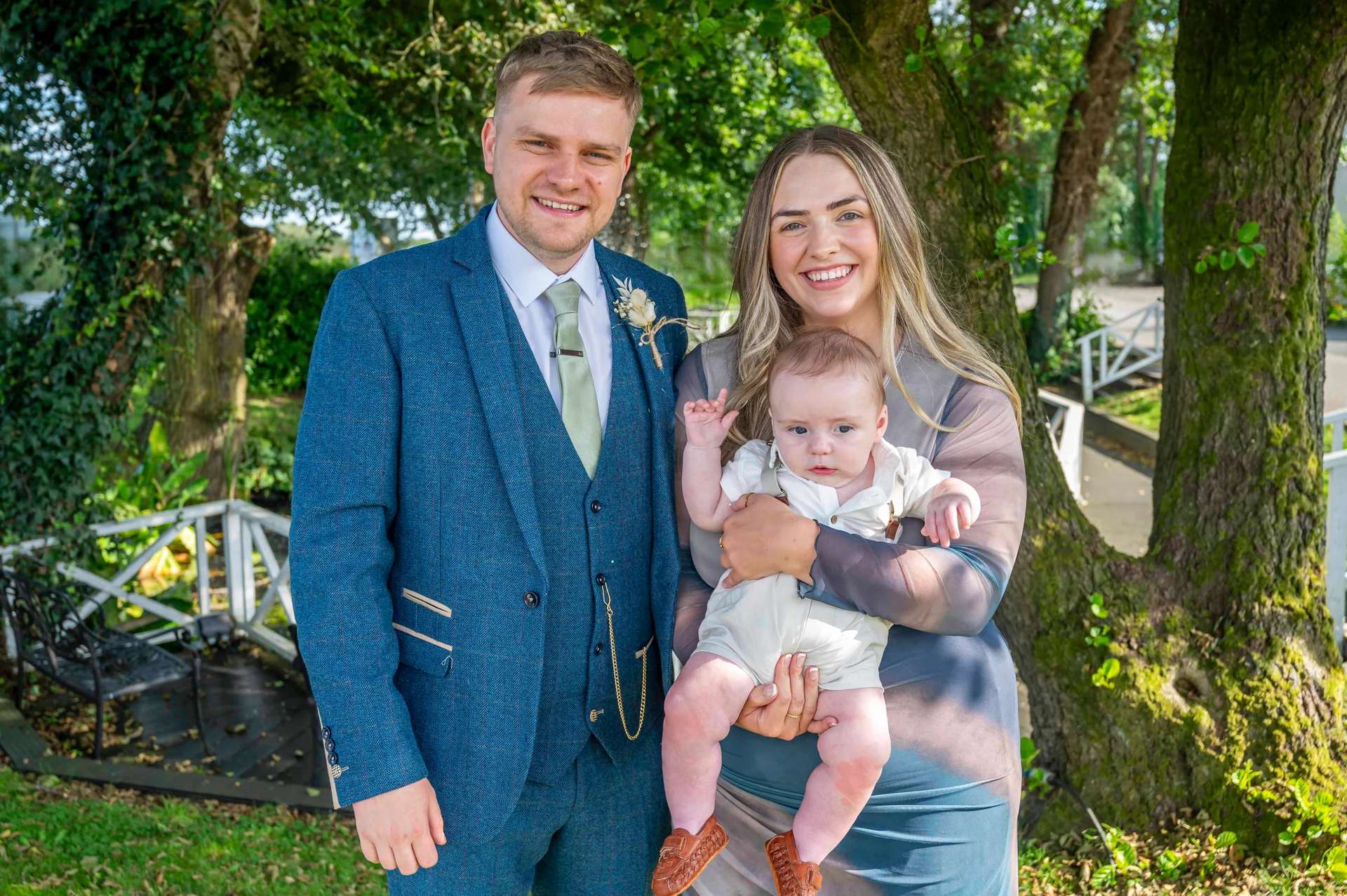 A smiling couple standing outdoors next to a mature tree, with the woman holding a baby who is waving to the camera.