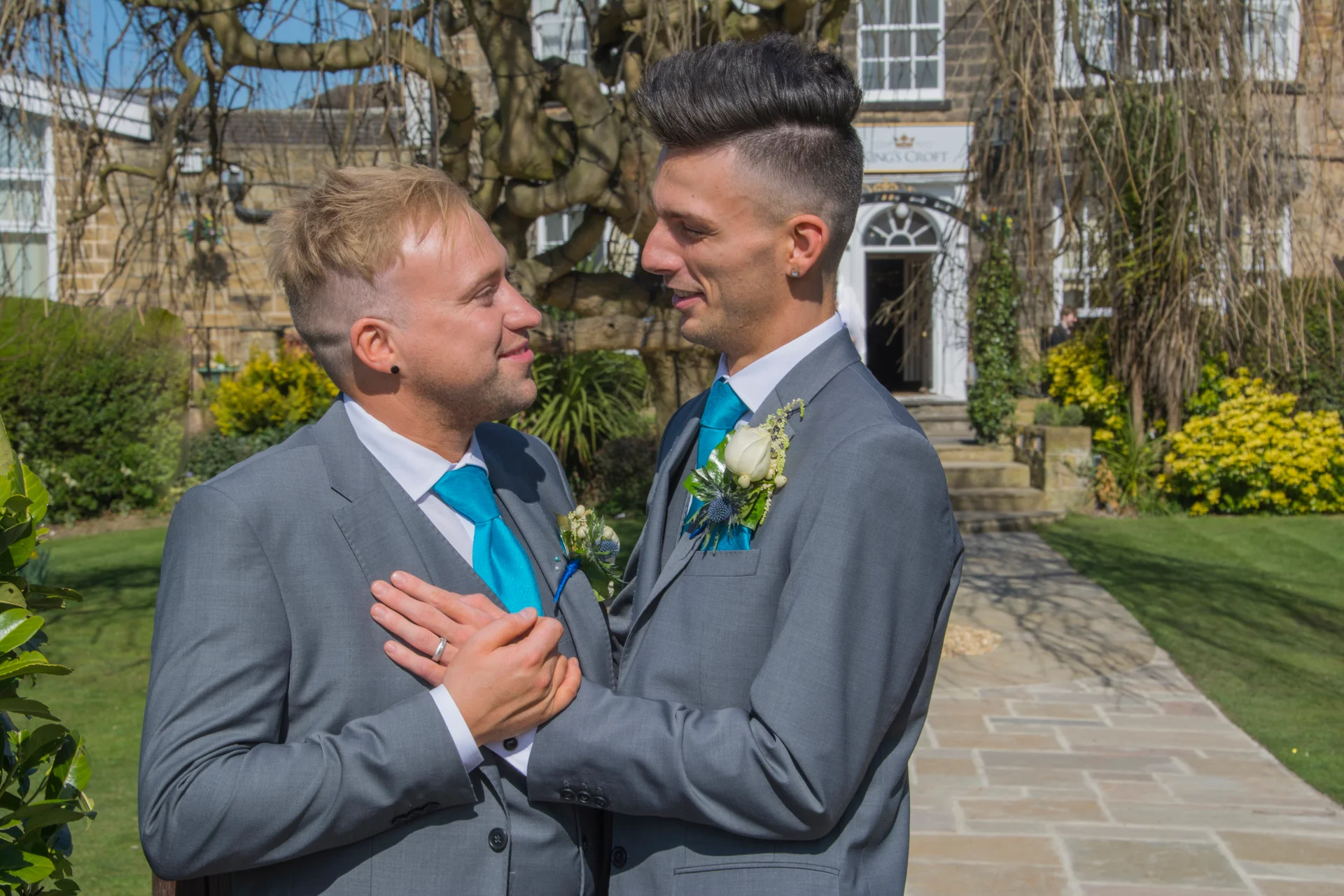 Two grooms in matching grey suits and bright turquoise ties share a tender, smiling moment holding hands outside a country house venue.