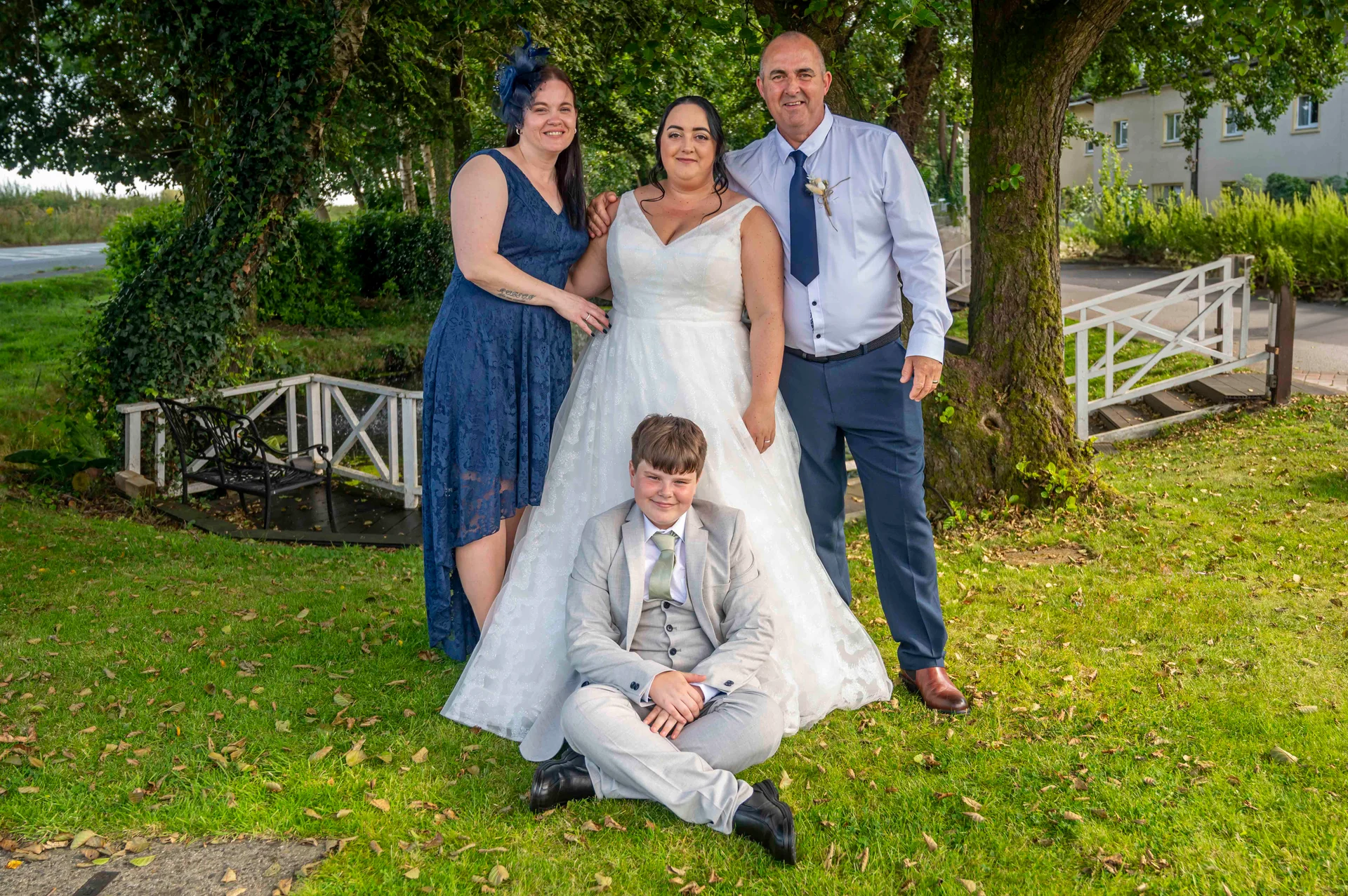 A smiling family group poses outdoors on a grassy lawn beside a large tree, featuring a bride in an a line tulle gown, a young page boy sitting cross legged in a grey suit, and guests in a blue dress and a grey suit.