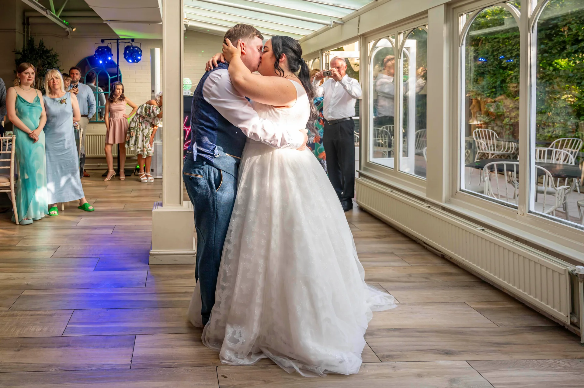Newlyweds share their first dance inside a bright conservatory while wedding guests look on and take photos.