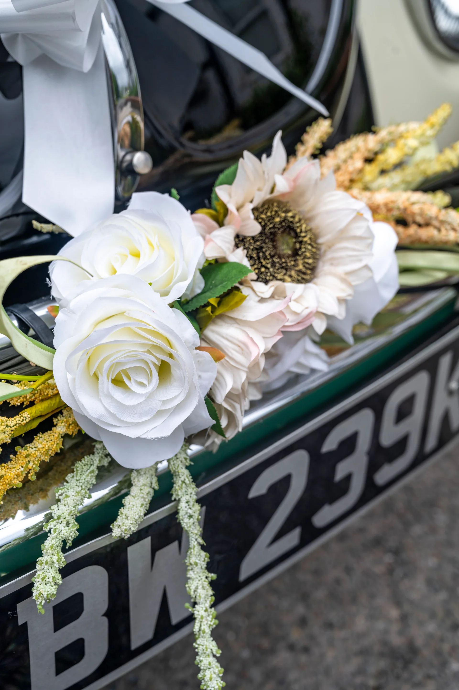 Close-up of a wedding car decorated with white roses, a sunflower, and trailing ribbons attached to the front bumper.