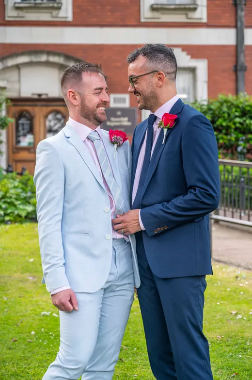 Two grooms in a light blue suit and a navy suit smile warmly at each other outside a red brick building on their wedding day.