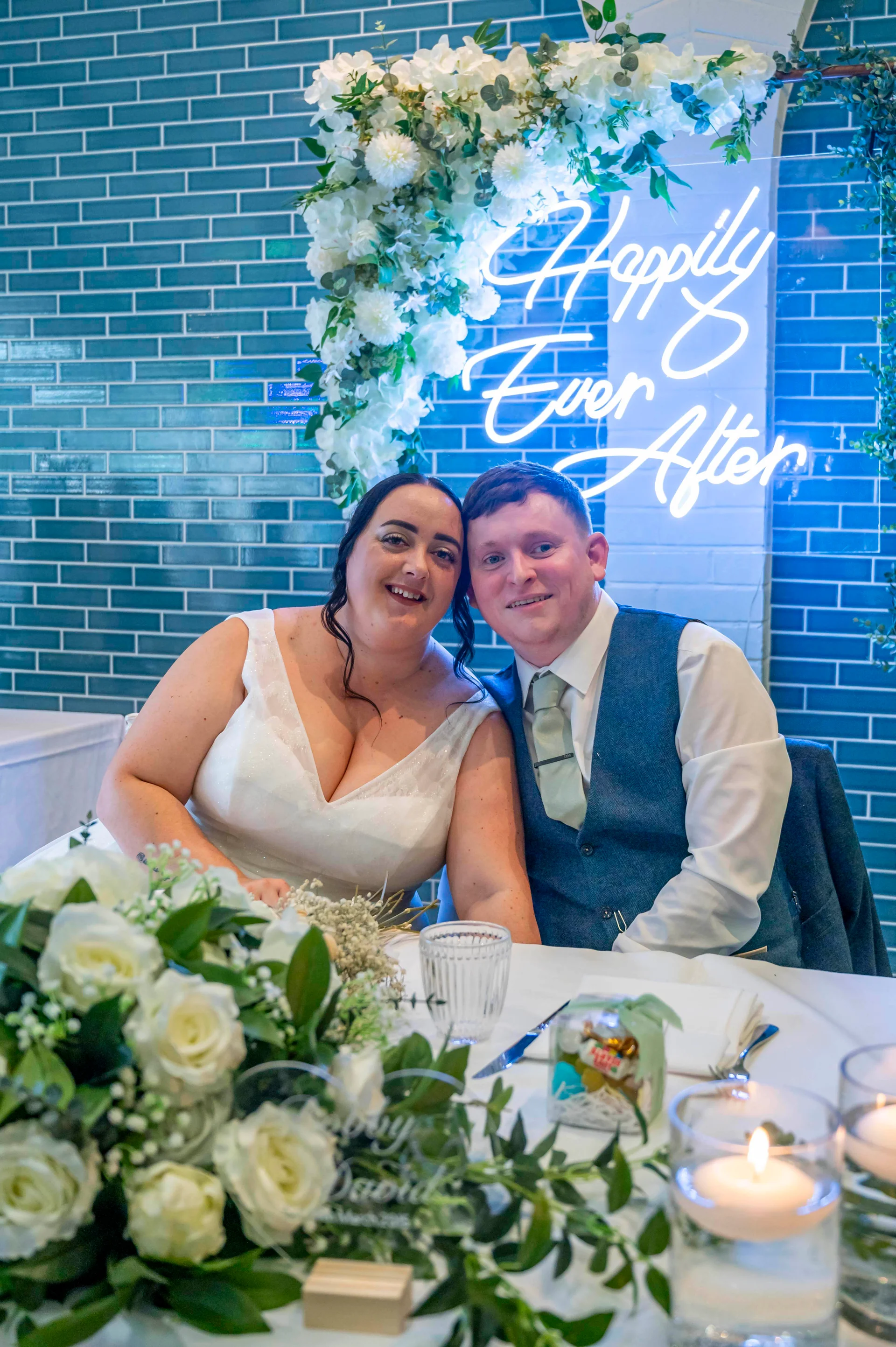 A smiling bride and groom sit together at the top table, framed by a blue tiled wall, white floral installations, and a glowing neon sign.